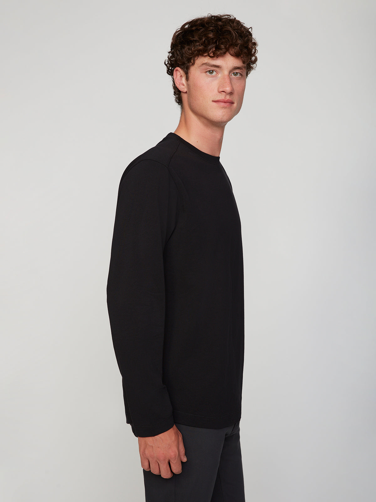 A young man with short curly brown hair stands sideways, wearing the PROJEK RAW MENS LONG SLEEVE CREW NECK paired with dark pants, against a plain light gray background.