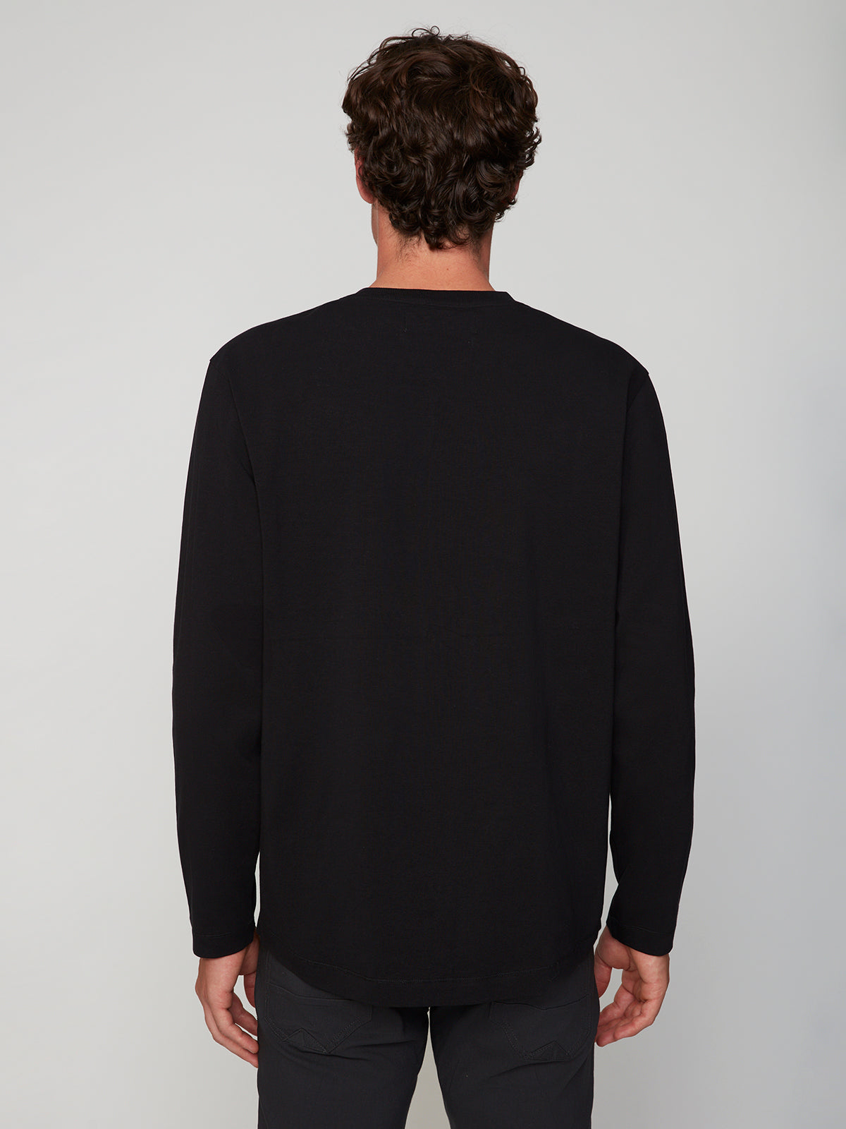 Seen from behind, a person with short curly brown hair wears a PROJEK RAW MENS LONG SLEEVE CREW NECK black shirt and dark pants, standing against a plain light background.