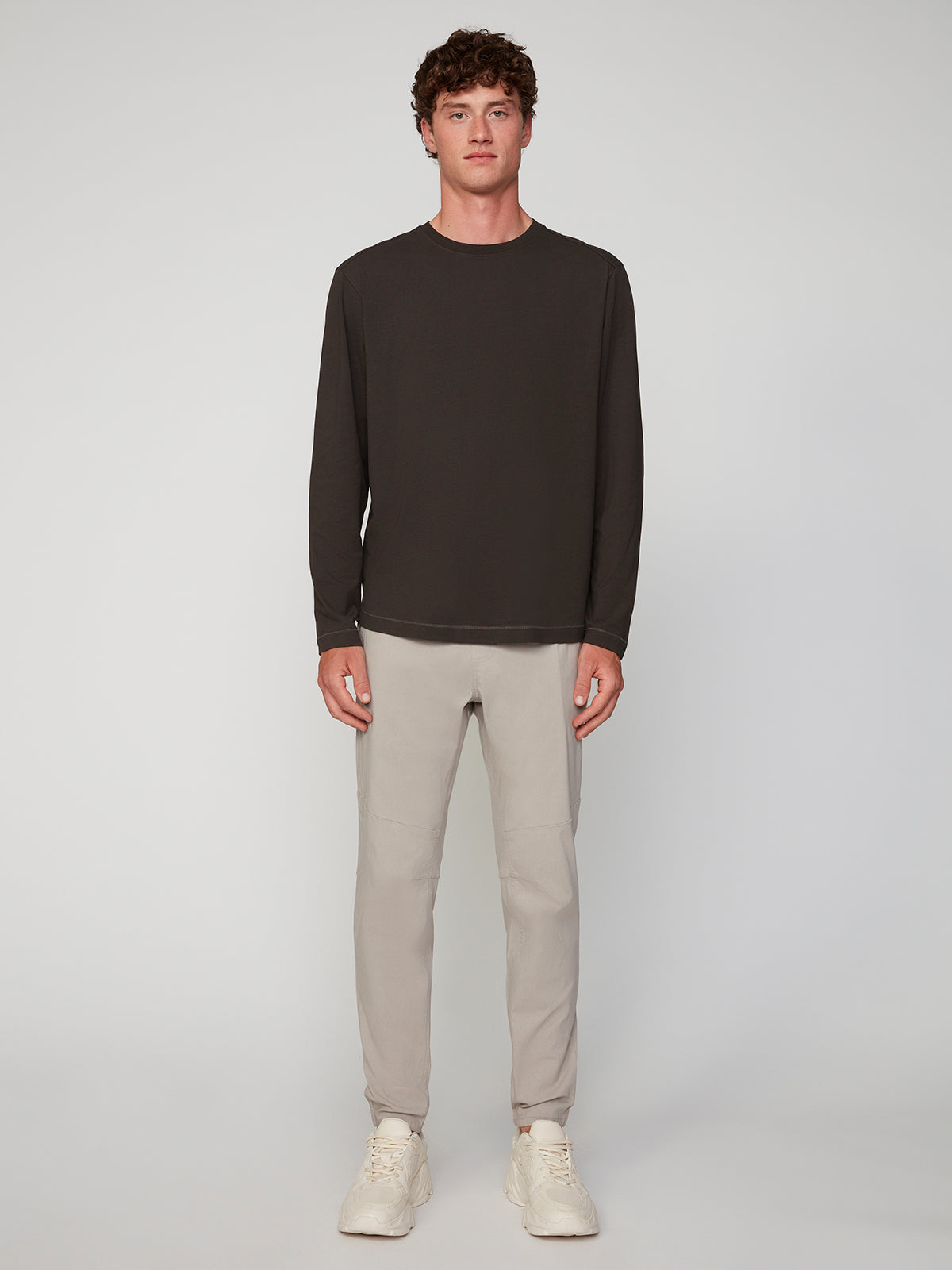 A man stands facing forward against a plain light background, wearing the PROJEK RAW MENS LONG SLEEVE CREW NECK, light gray pants, and white sneakers.