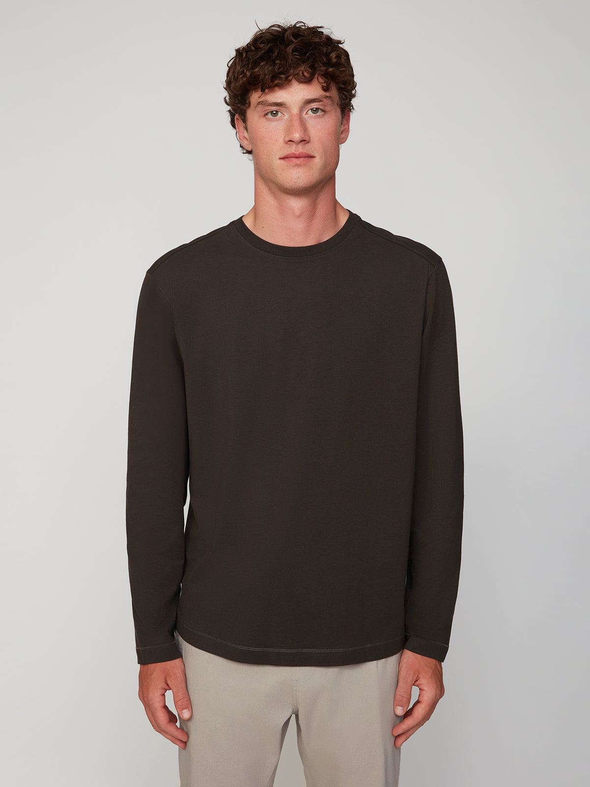 A young man with curly brown hair wears a PROJEK RAW MENS LONG SLEEVE CREW NECK in dark brown and light beige pants, standing facing forward against a plain light gray background.