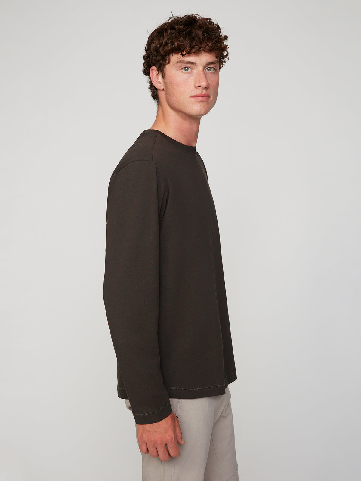 A young man with curly brown hair stands sideways, facing left, wearing a PROJEK RAW MENS LONG SLEEVE CREW NECK in dark brown and light pants, with a neutral expression against a plain light background.