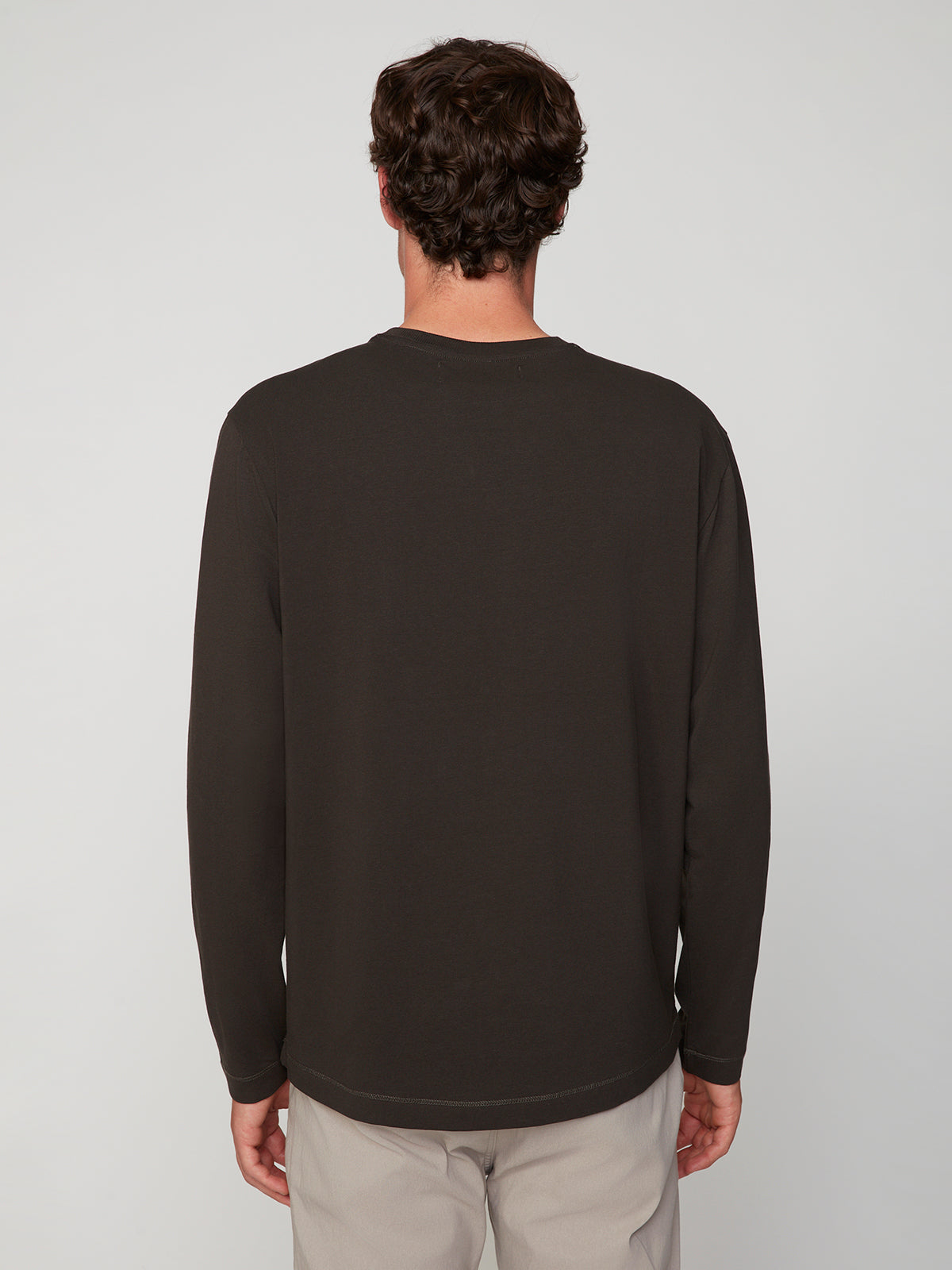 A man with short, curly brown hair stands with his back to the camera in a MENS LONG SLEEVE CREW NECK by PROJEK RAW—a versatile stretch cotton layer—paired with light pants against a plain background.