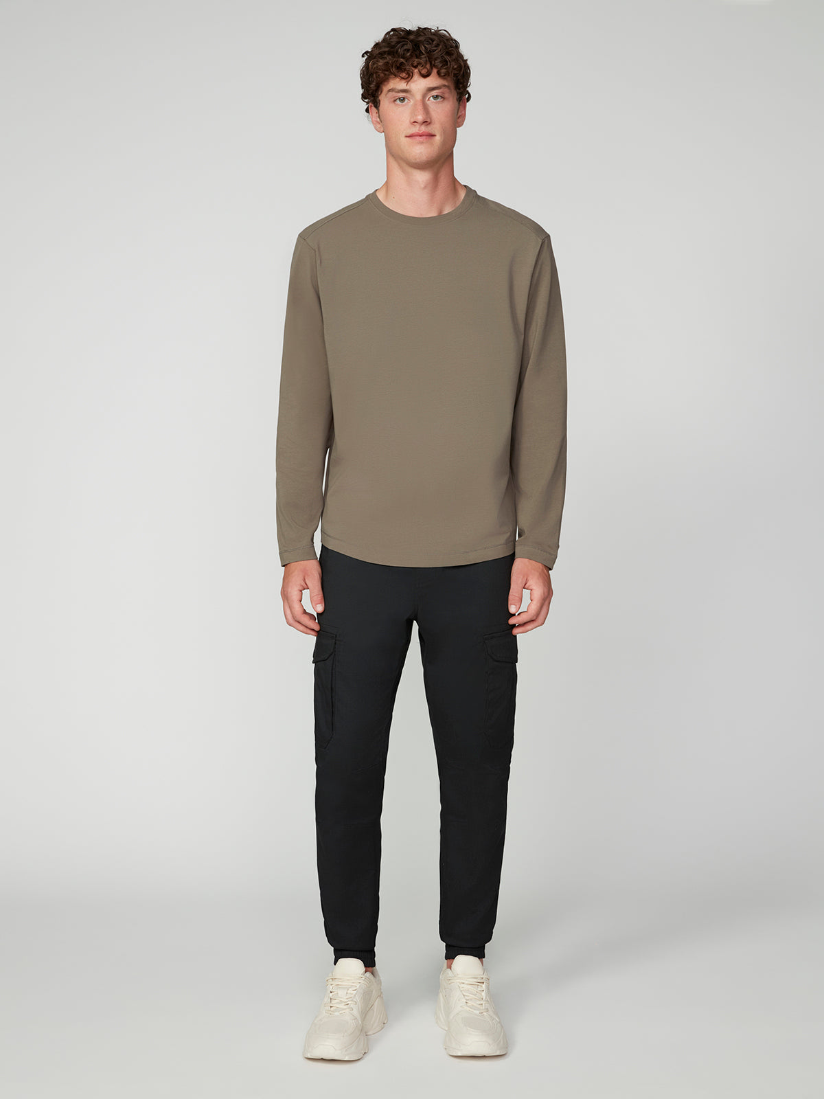 A person stands facing forward against a plain light gray background, wearing the PROJEK RAW MENS LONG SLEEVE CREW NECK in olive green, paired with black cargo joggers and white sneakers.