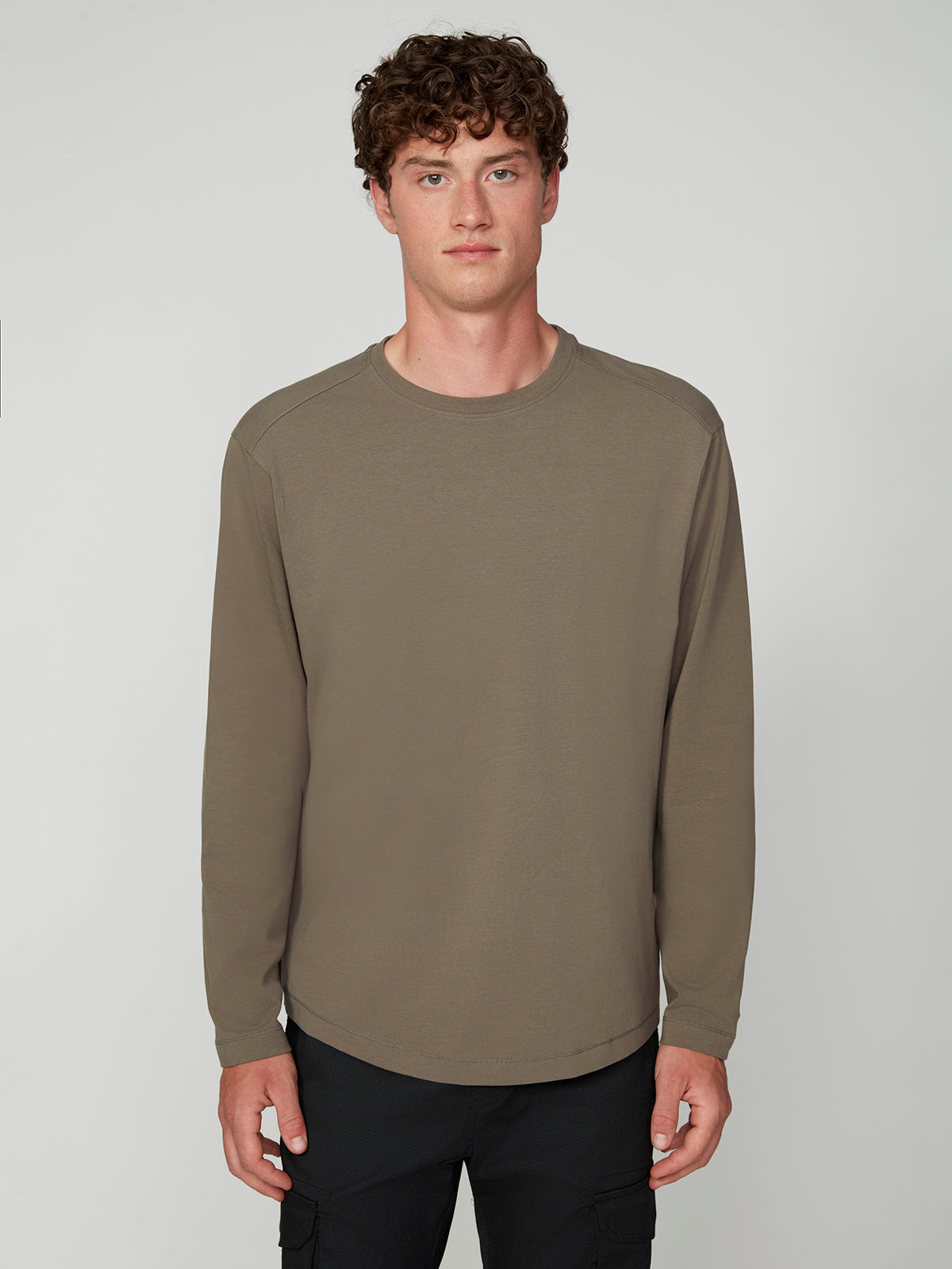 A young man with curly brown hair wears the PROJEK RAW MENS LONG SLEEVE CREW NECK in olive green stretch cotton jersey and black pants, standing against a plain light gray background, facing forward with a neutral expression.