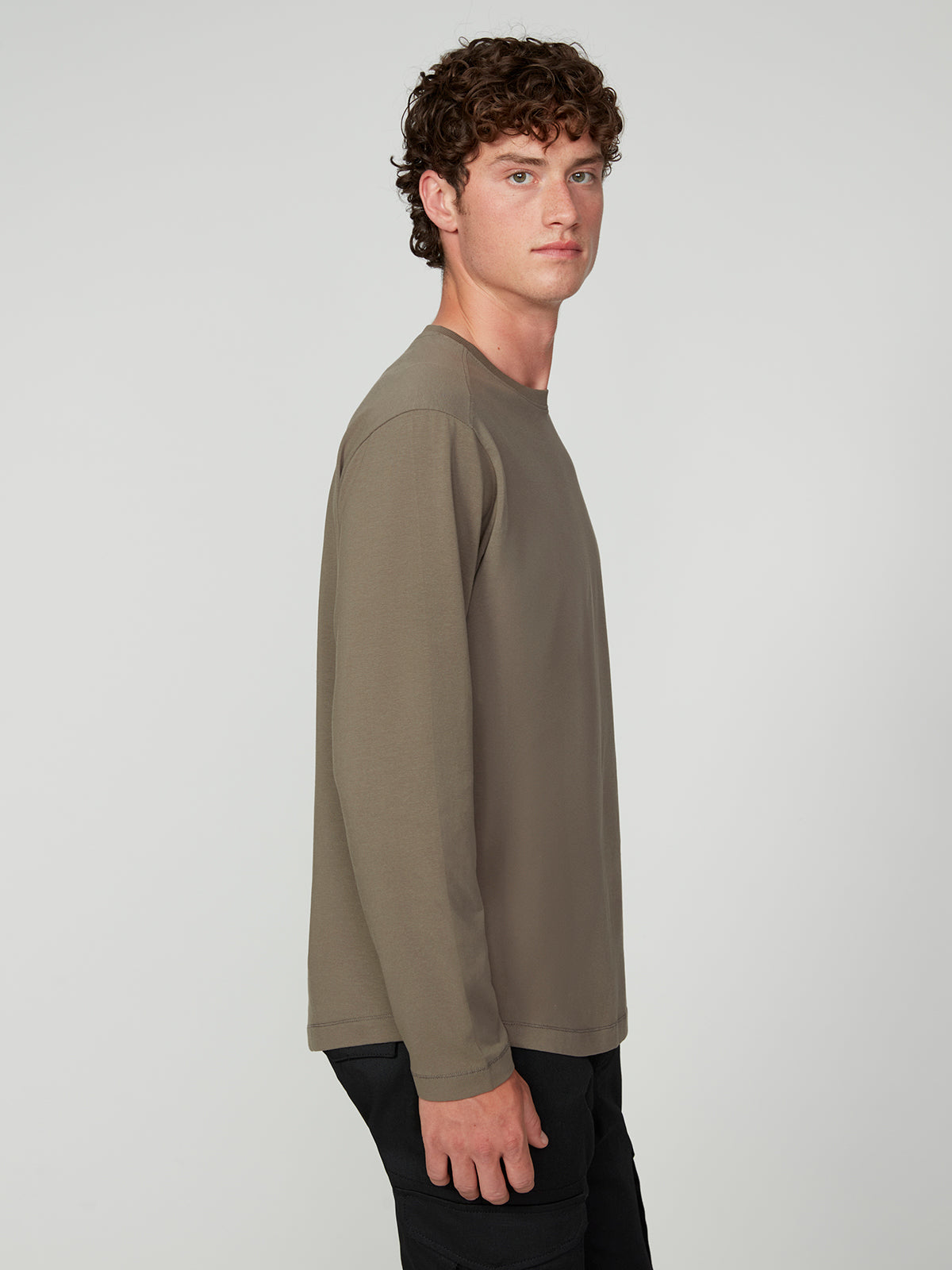 A young man with curly brown hair stands in profile against a light gray background, wearing a taupe MENS LONG SLEEVE CREW NECK by PROJEK RAW and black pants.