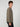 A young man with curly brown hair stands in profile against a light gray background, wearing a taupe MENS LONG SLEEVE CREW NECK by PROJEK RAW and black pants.