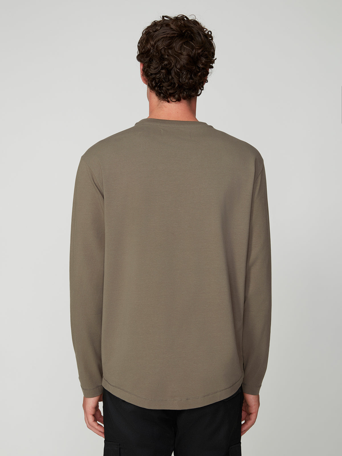 A man with short curly brown hair faces away from the camera, wearing the PROJEK RAW MENS LONG SLEEVE CREW NECK and black pants against a plain light gray background. Ideal for mens layering.