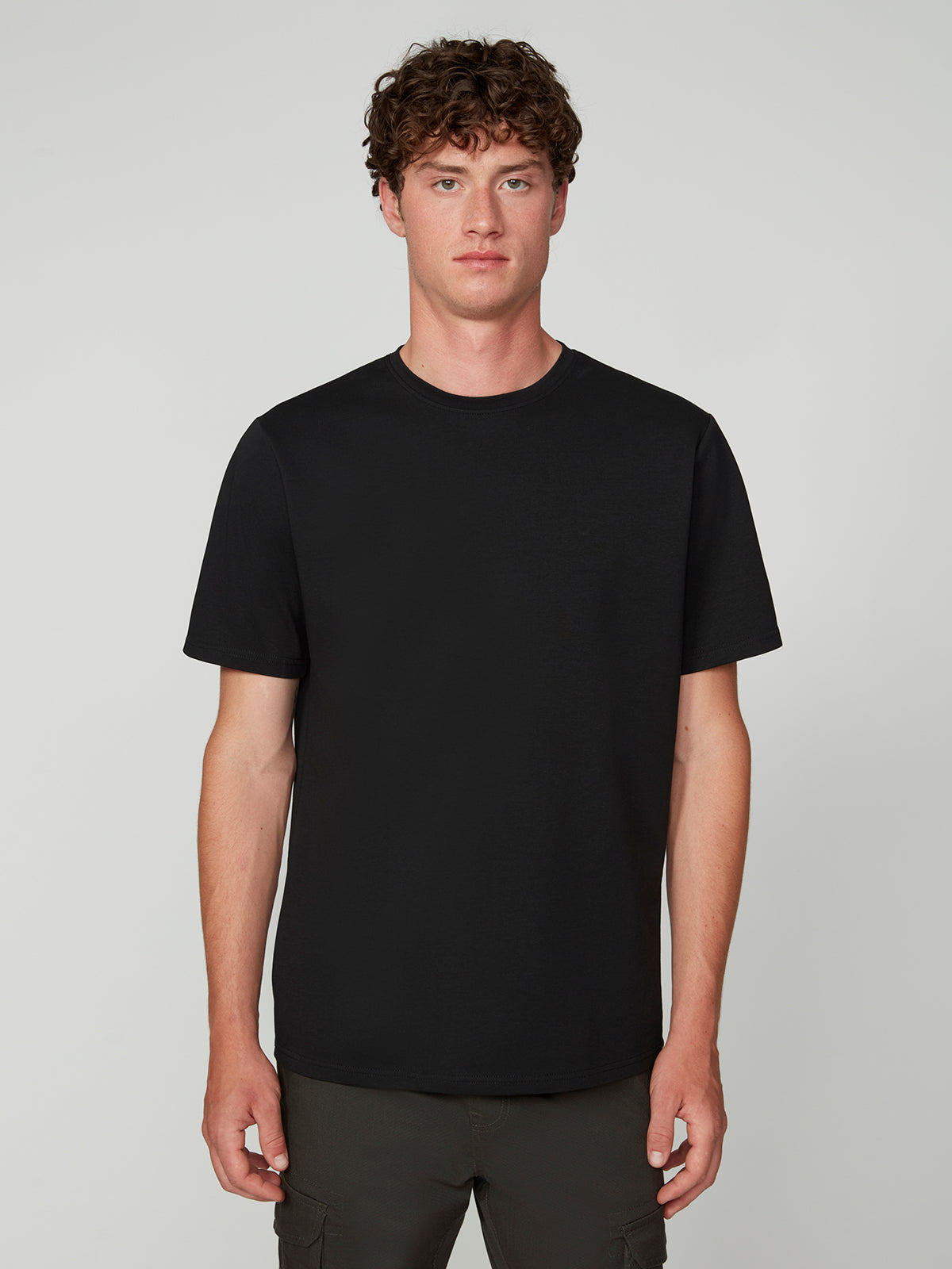 A young man with curly brown hair stands facing forward against a light gray background, showcasing the PROJEK RAW MENS SHORT SLEEVE SCUBA JERSEY RELAXED FIT TSHIRT in black and dark pants for a modern casual look.