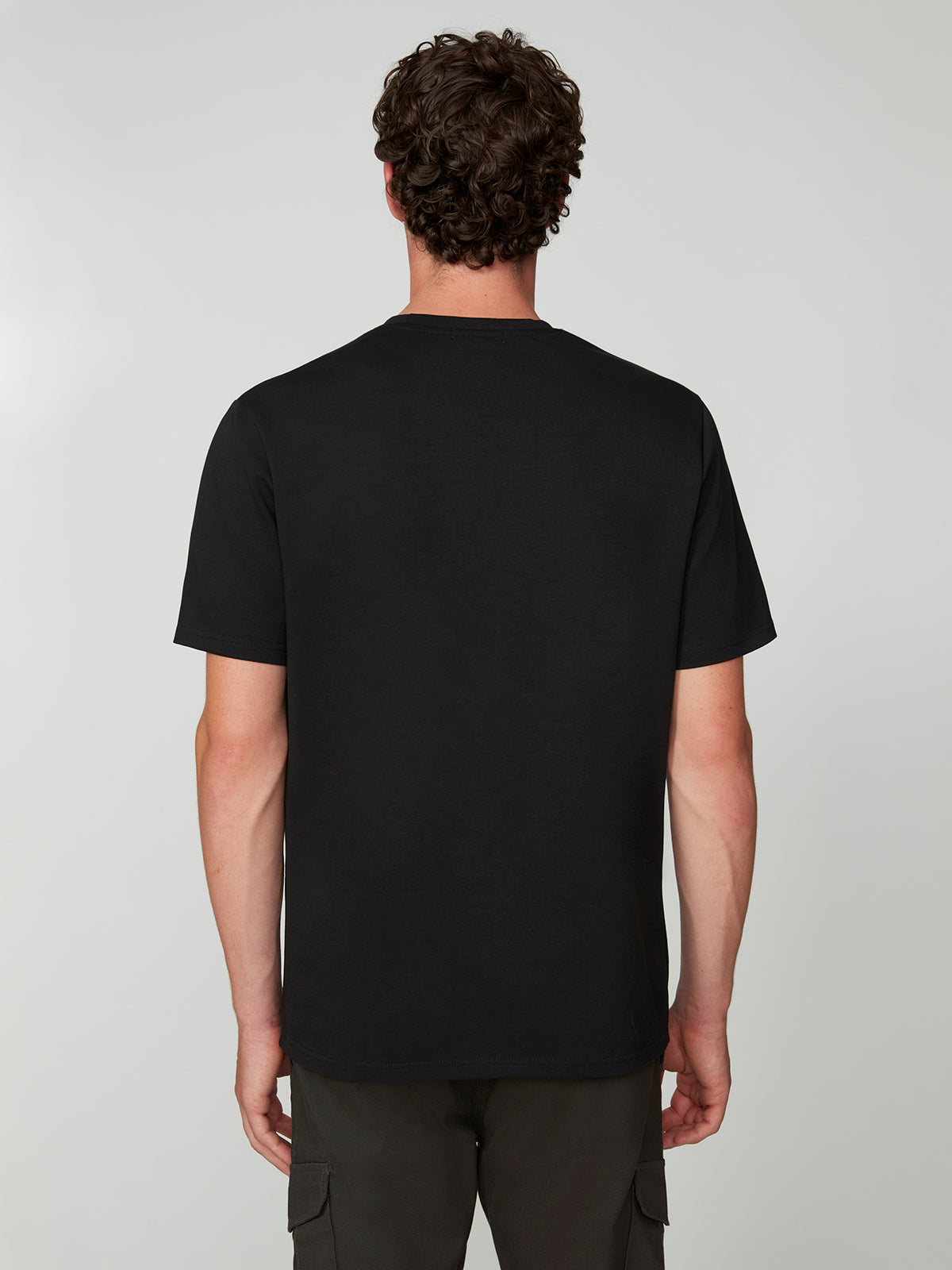 A man with curly brown hair is seen from behind, wearing a PROJEK RAW MENS SHORT SLEEVE SCUBA JERSEY RELAXED FIT TSHIRT in black and dark cargo pants, standing against a light gray background.