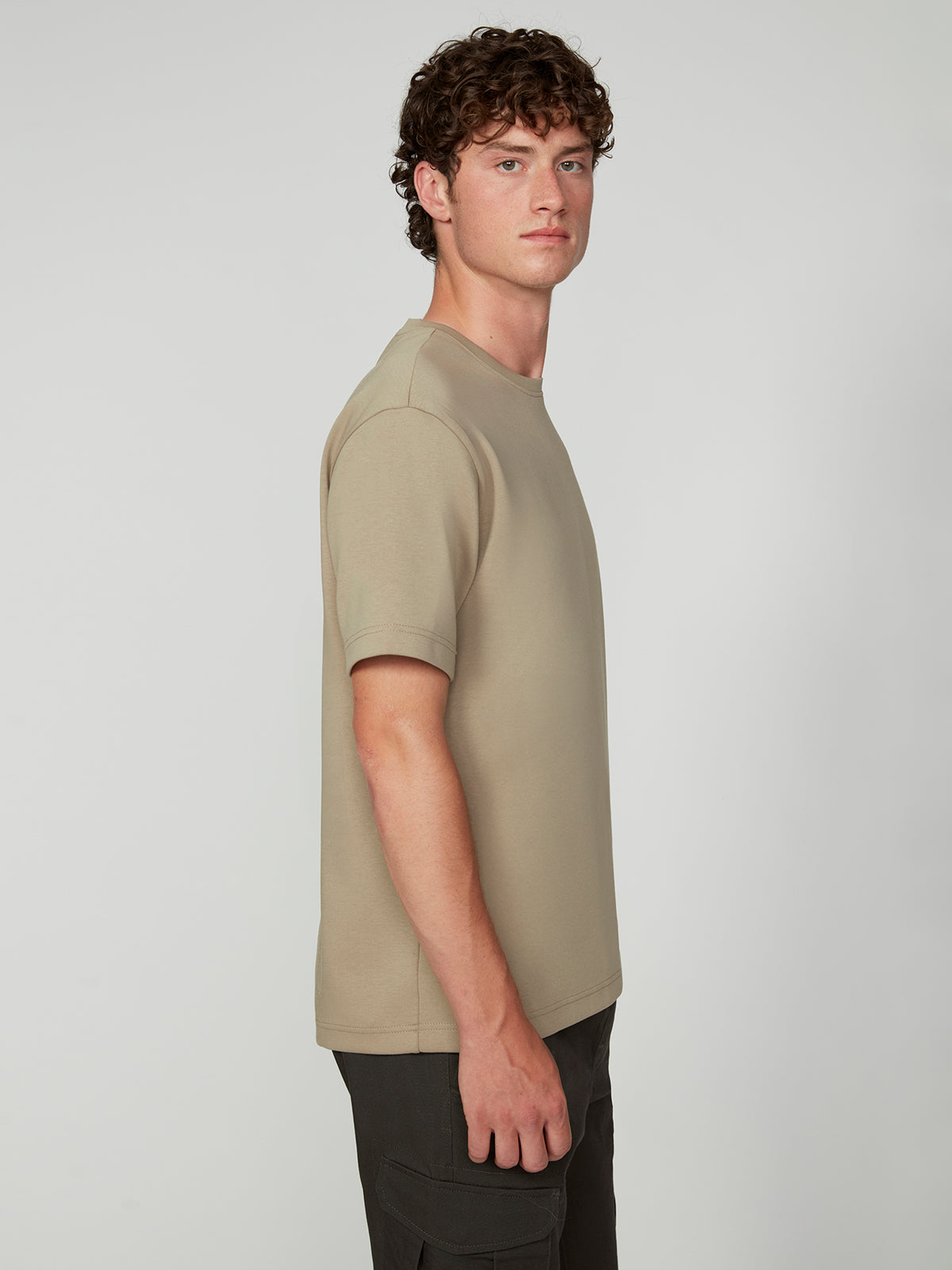 A young man with curly brown hair, wearing a beige PROJEK RAW MENS SHORT SLEEVE SCUBA JERSEY RELAXED FIT TSHIRT and dark pants, stands sideways against a plain light gray background.