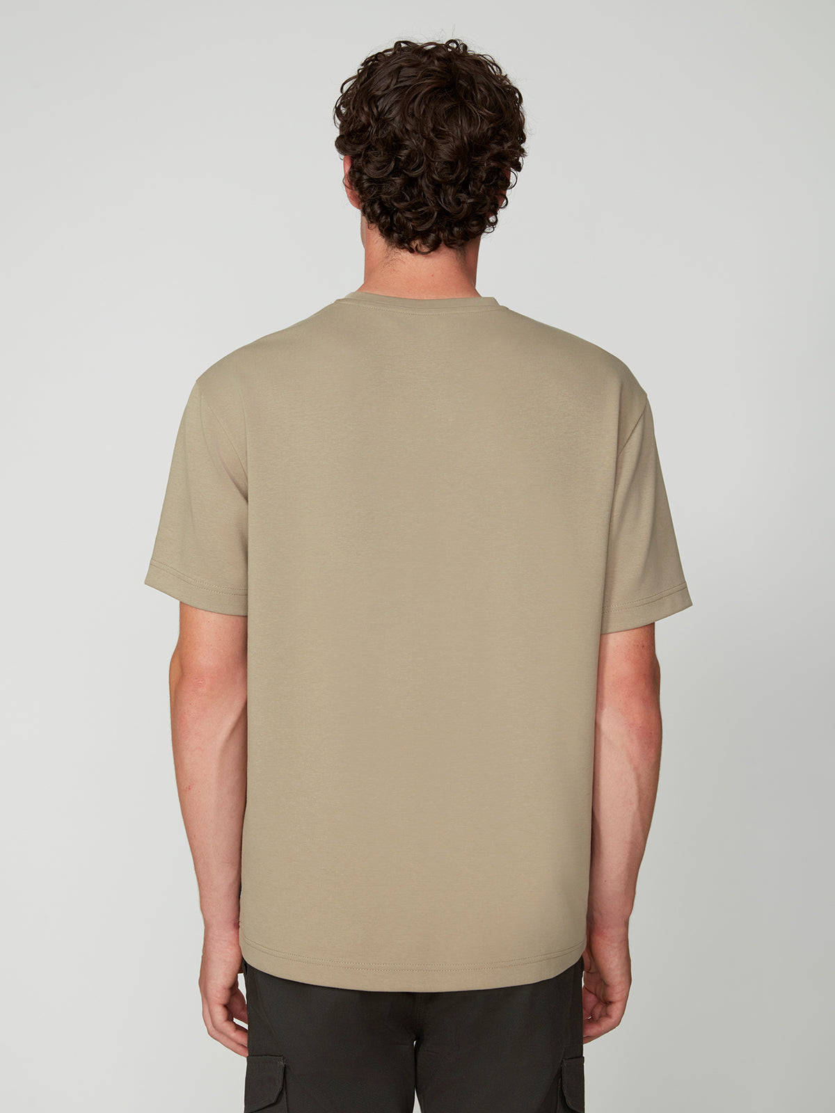 A man with curly brown hair stands facing away from the camera, wearing a PROJEK RAW MENS SHORT SLEEVE SCUBA JERSEY RELAXED FIT TSHIRT in beige and dark shorts, styled casually against a neutral background.