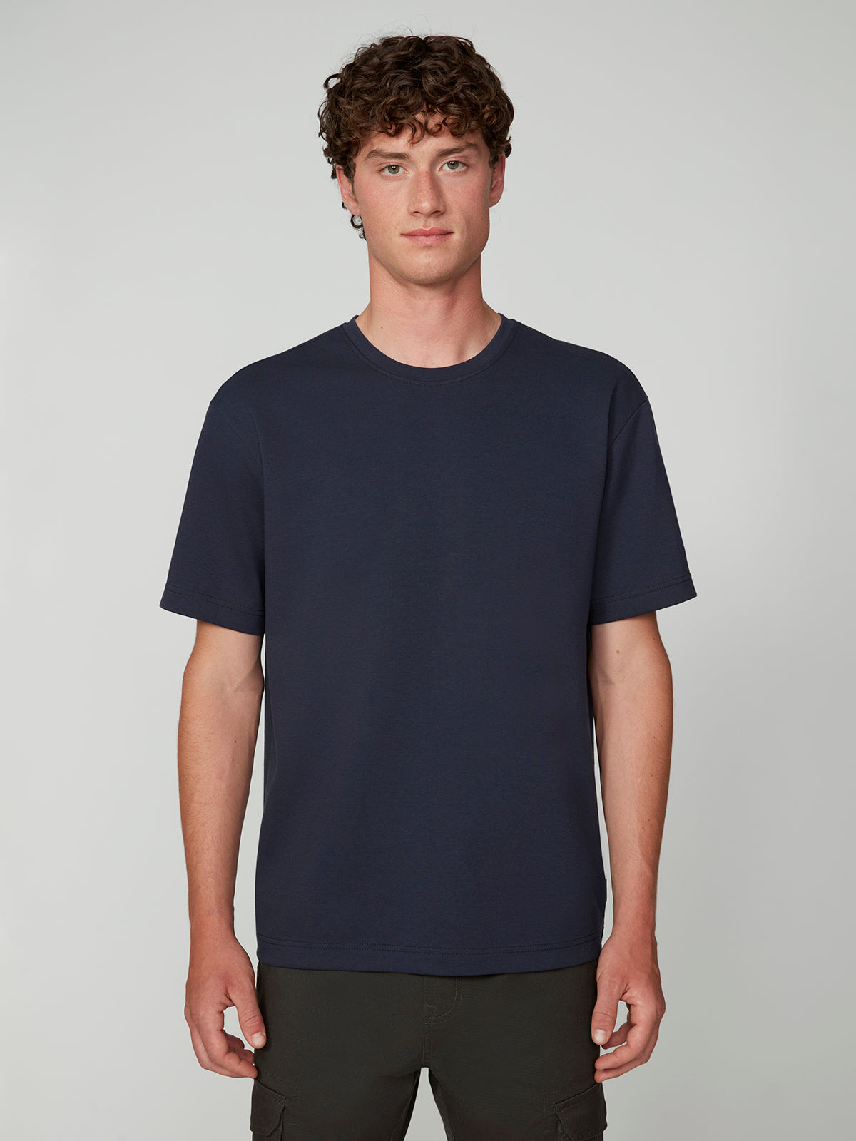 A young man with curly brown hair faces the camera, wearing a PROJEK RAW MENS SHORT SLEEVE SCUBA JERSEY RELAXED FIT TSHIRT in navy blue and dark pants against a neutral light gray background—perfect for casual wear.