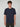 A young man with curly brown hair faces the camera, wearing a PROJEK RAW MENS SHORT SLEEVE SCUBA JERSEY RELAXED FIT TSHIRT in navy blue and dark pants against a neutral light gray background—perfect for casual wear.