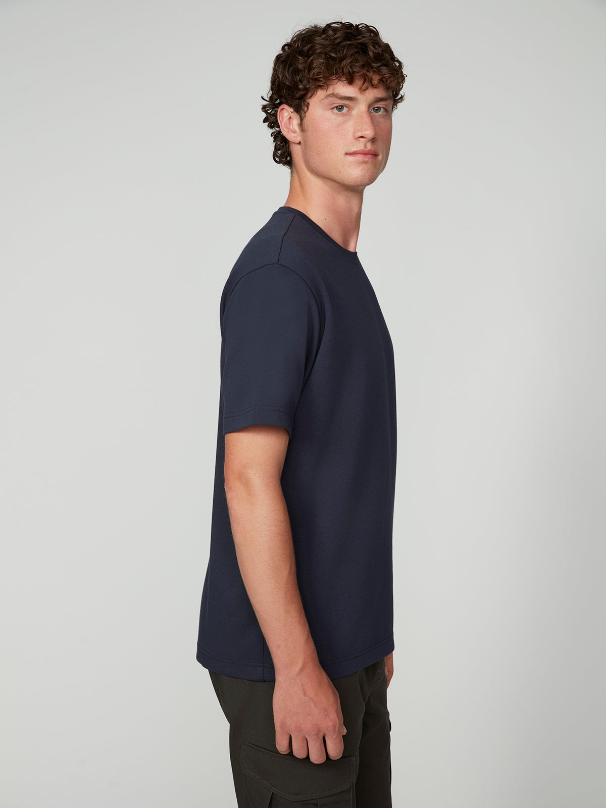 A young man with short curly brown hair models the PROJEK RAW MENS SHORT SLEEVE SCUBA JERSEY RELAXED FIT TSHIRT in navy blue and black pants, standing sideways against a plain light gray background, ideal for casual wear.
