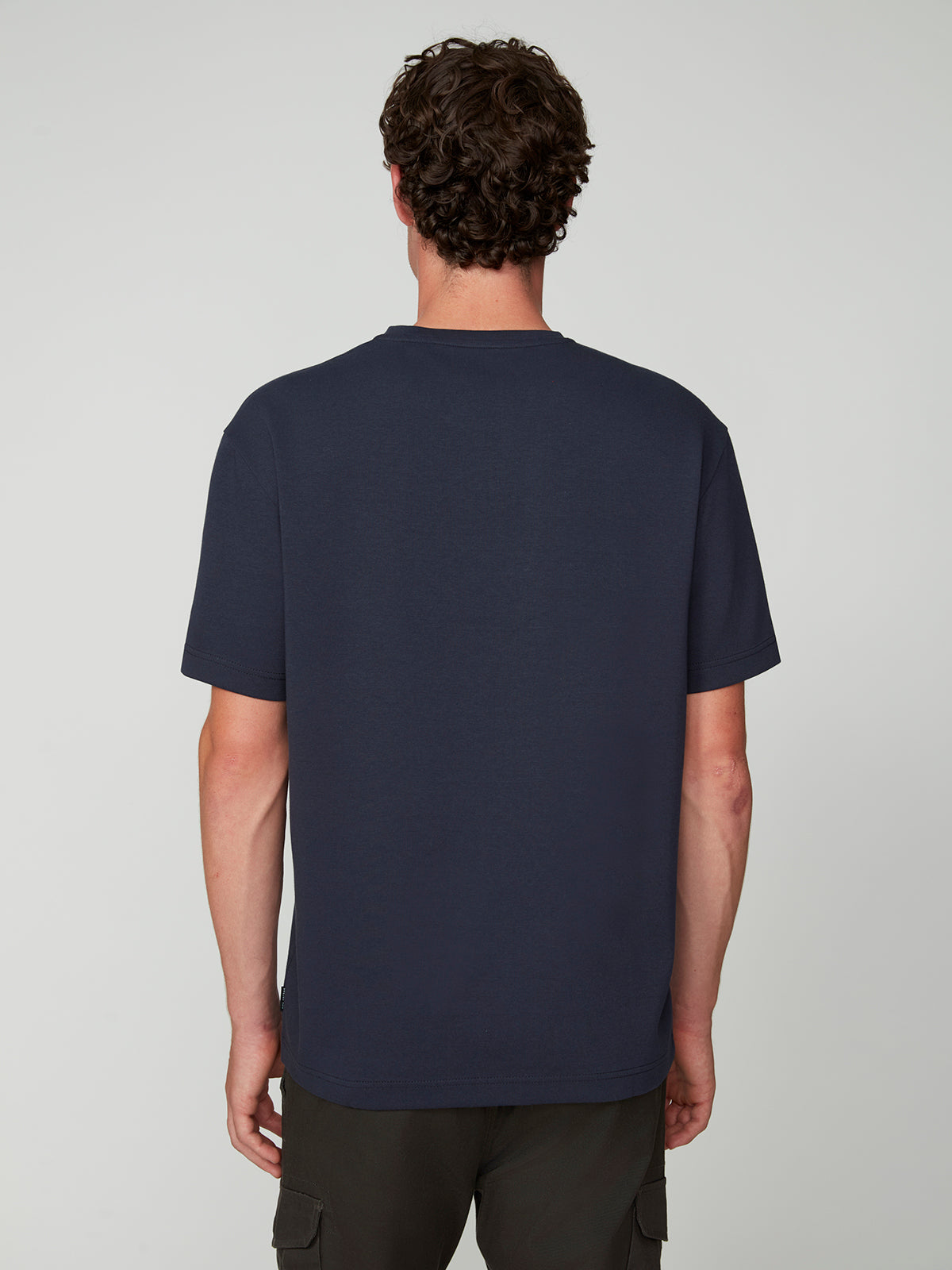 A person with short, curly brown hair stands with their back to the camera, wearing a PROJEK RAW MENS SHORT SLEEVE SCUBA JERSEY RELAXED FIT TSHIRT in navy blue and dark cargo pants, set against a light gray background.