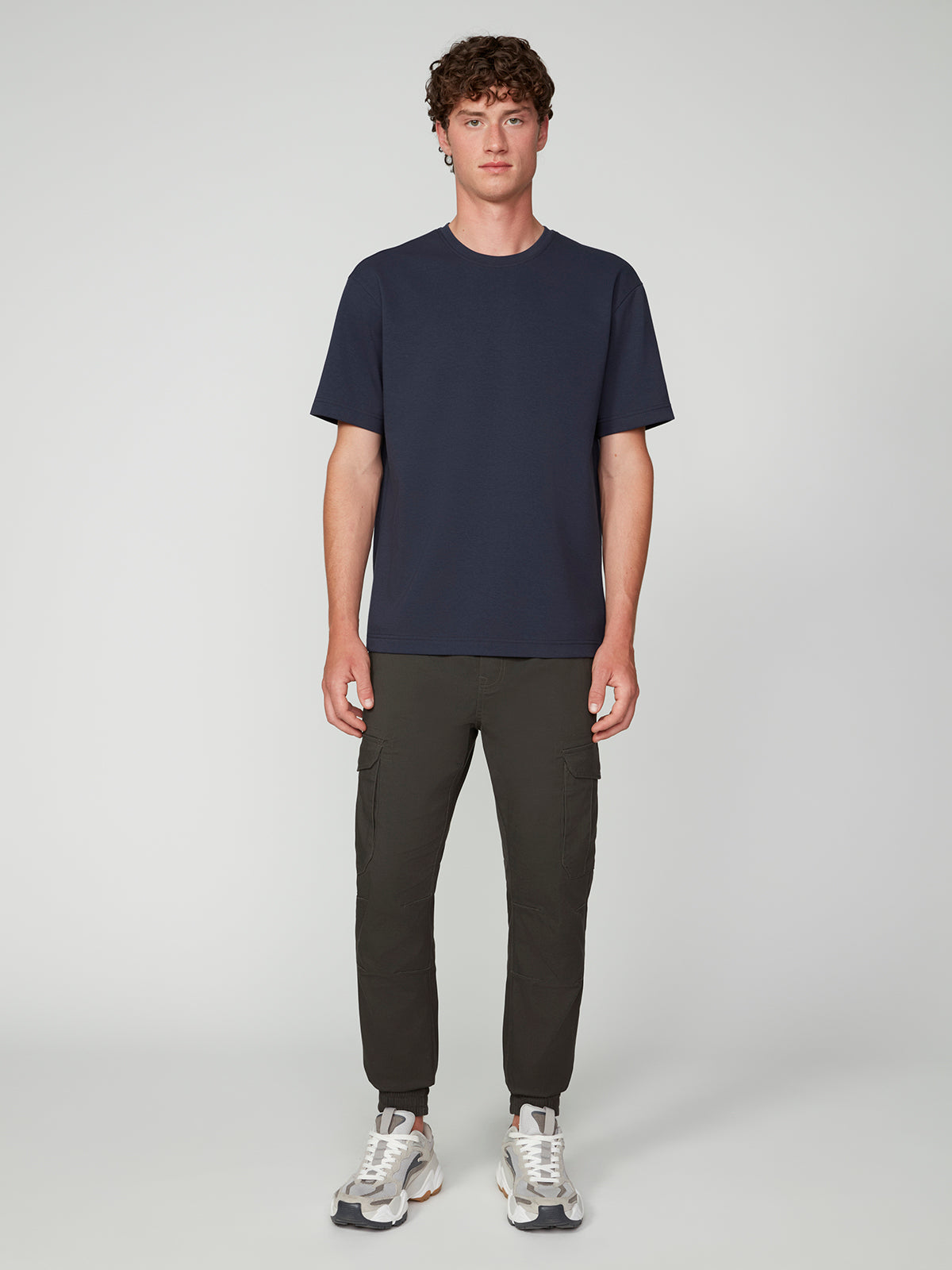 A man with short curly hair stands against a light gray background, wearing a PROJEK RAW MENS SHORT SLEEVE SCUBA JERSEY RELAXED FIT TSHIRT, olive green cargo pants, and white sneakers for a perfect casual look.