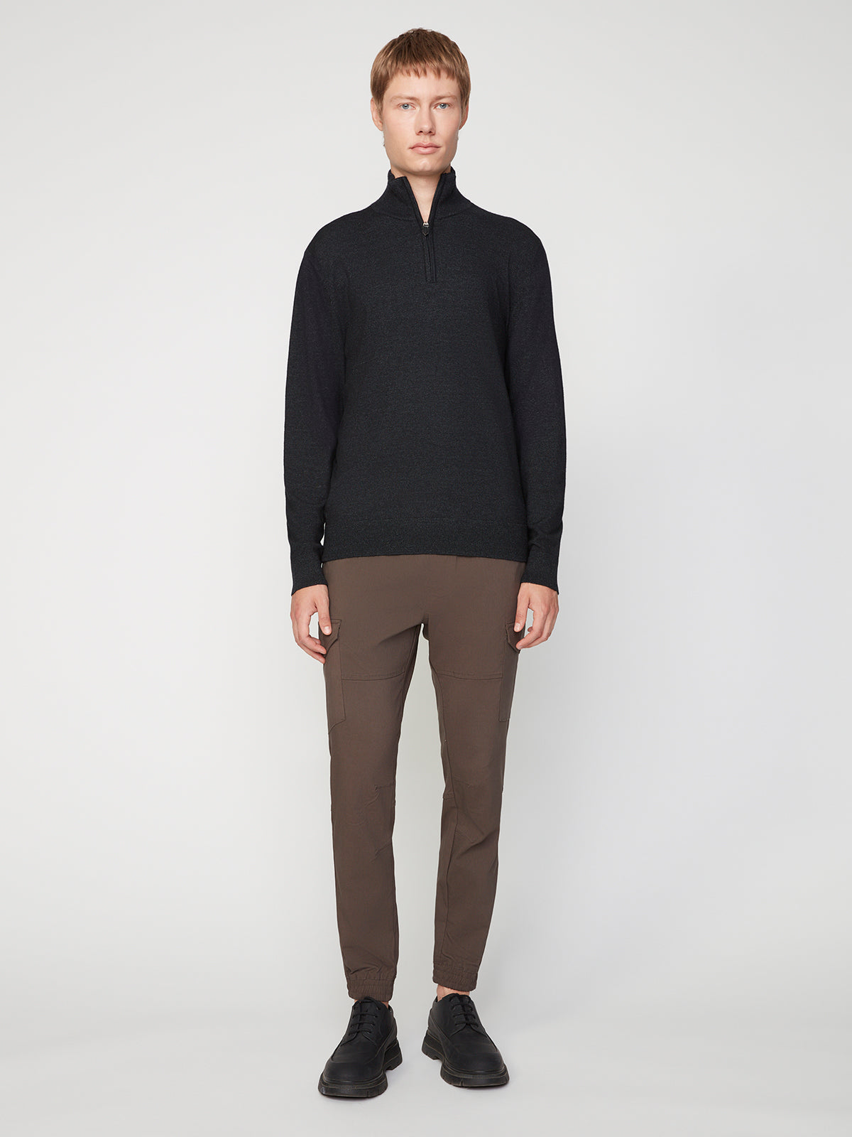 A person with short light brown hair wearing a PROJEK RAW MENS 1/4-ZIP CASHMERE TOUCH SWEATER, brown slim-fit pants, and black shoes stands facing forward against a plain light gray background.