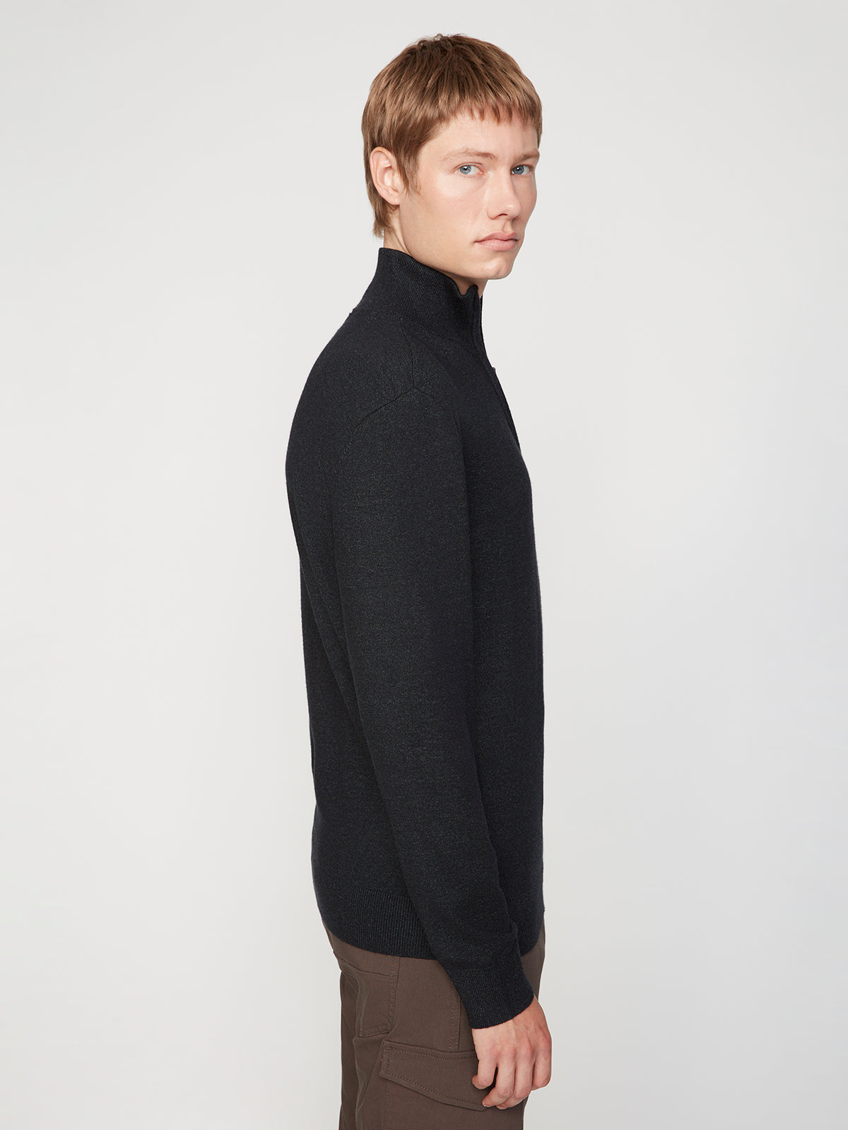 A person with short light brown hair, wearing the PROJEK RAW MENS 1/4-ZIP CASHMERE TOUCH SWEATER in black and brown pants, stands in profile against a plain white background.