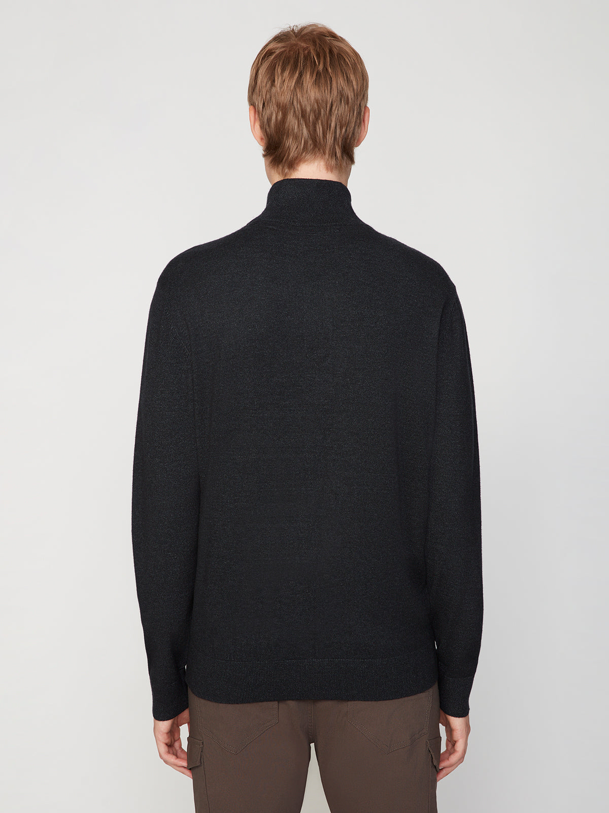 A person with light brown hair is shown from behind, wearing a black PROJEK RAW MENS 1/4-ZIP CASHMERE TOUCH SWEATER and brown pants, standing against a plain light gray background.