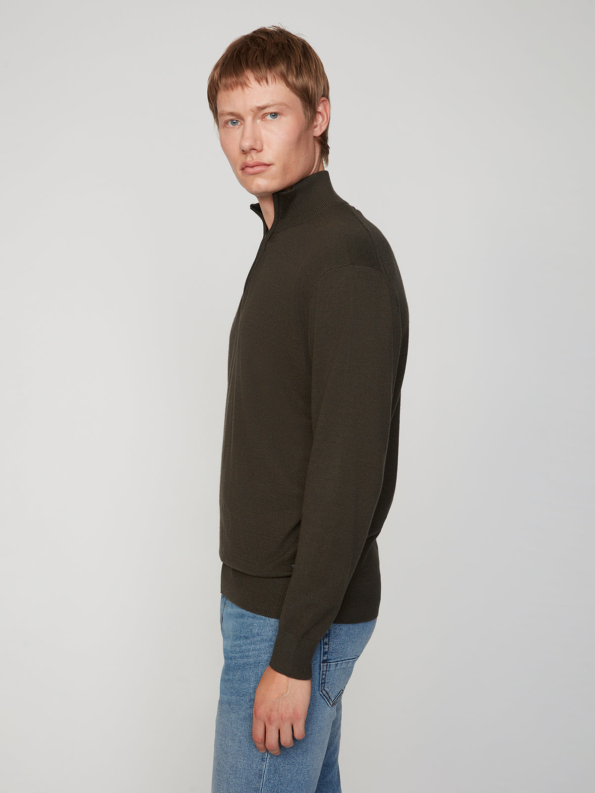 A young man with short auburn hair models the PROJEK RAW MENS 1/4-ZIP CASHMERE TOUCH SWEATER in dark olive and light blue jeans, standing sideways and looking at the camera against a plain light background.