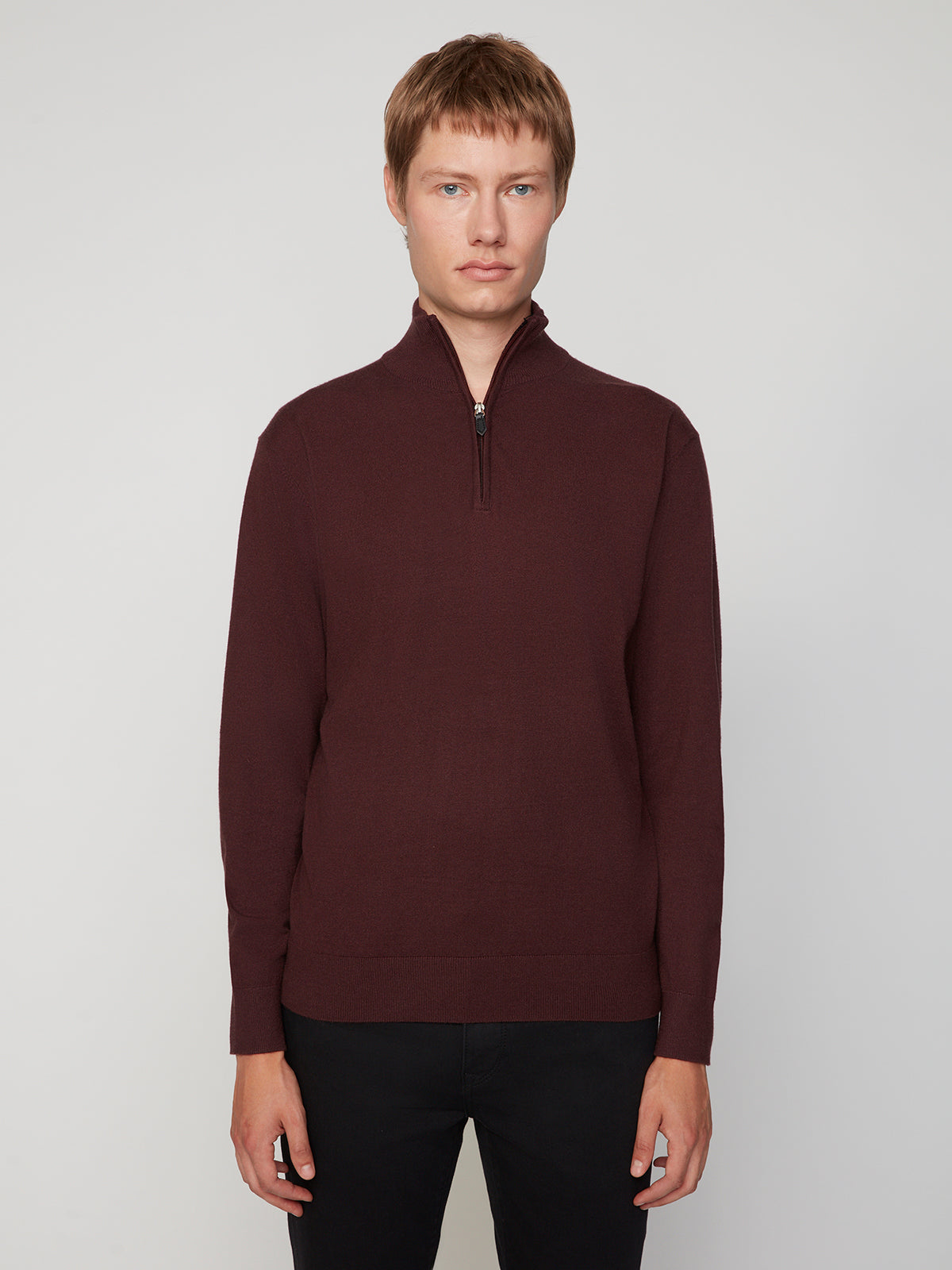 A person with short light brown hair wears a maroon MENS 1/4-ZIP CASHMERE TOUCH SWEATER by PROJEK RAW and black pants, standing against a plain light gray background, looking directly at the camera with a neutral expression.