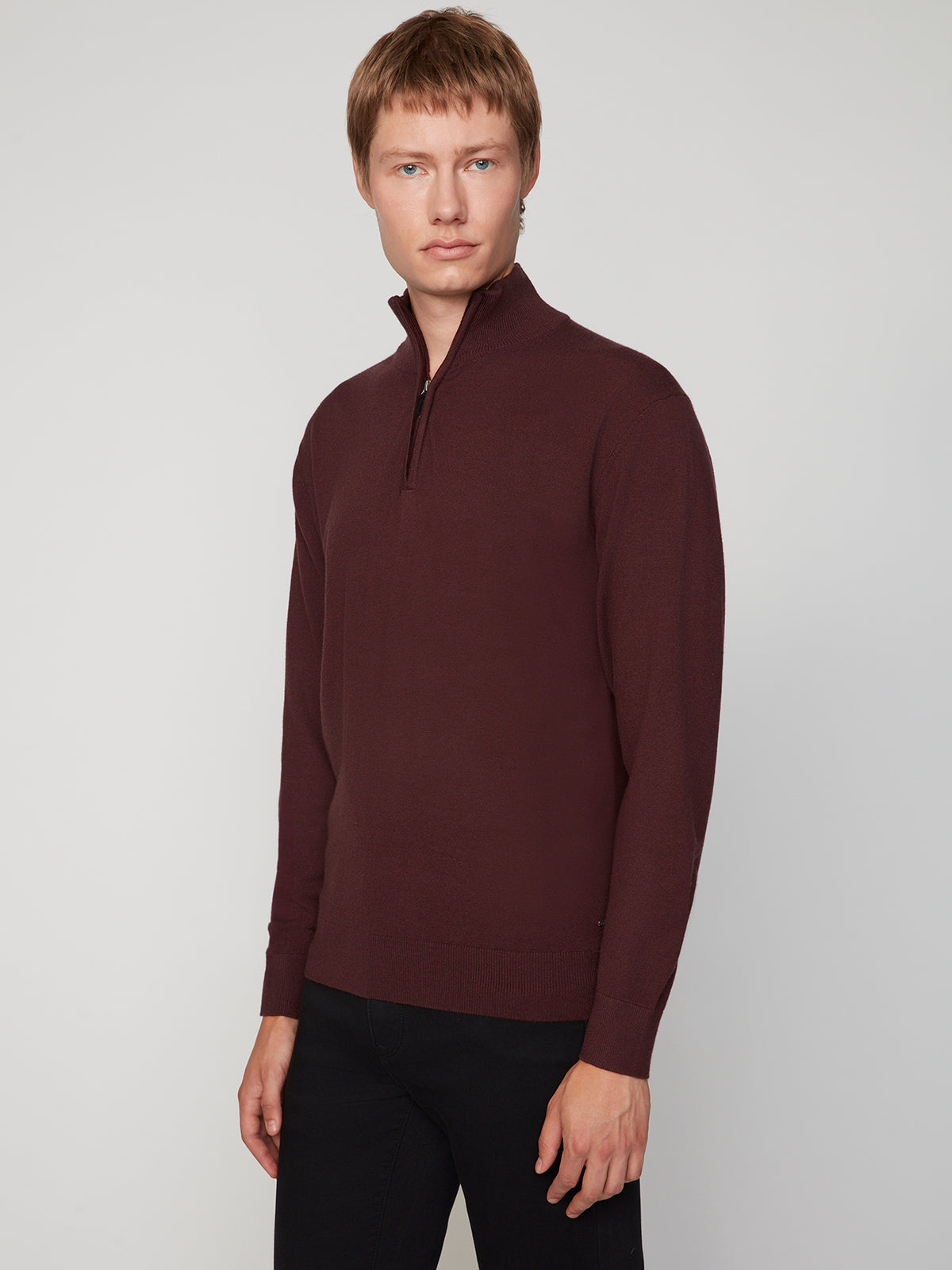 A man wears a maroon PROJEK RAW MENS 1/4-ZIP CASHMERE TOUCH SWEATER and black pants, standing against a plain light gray background, looking slightly to the side with a neutral expression.