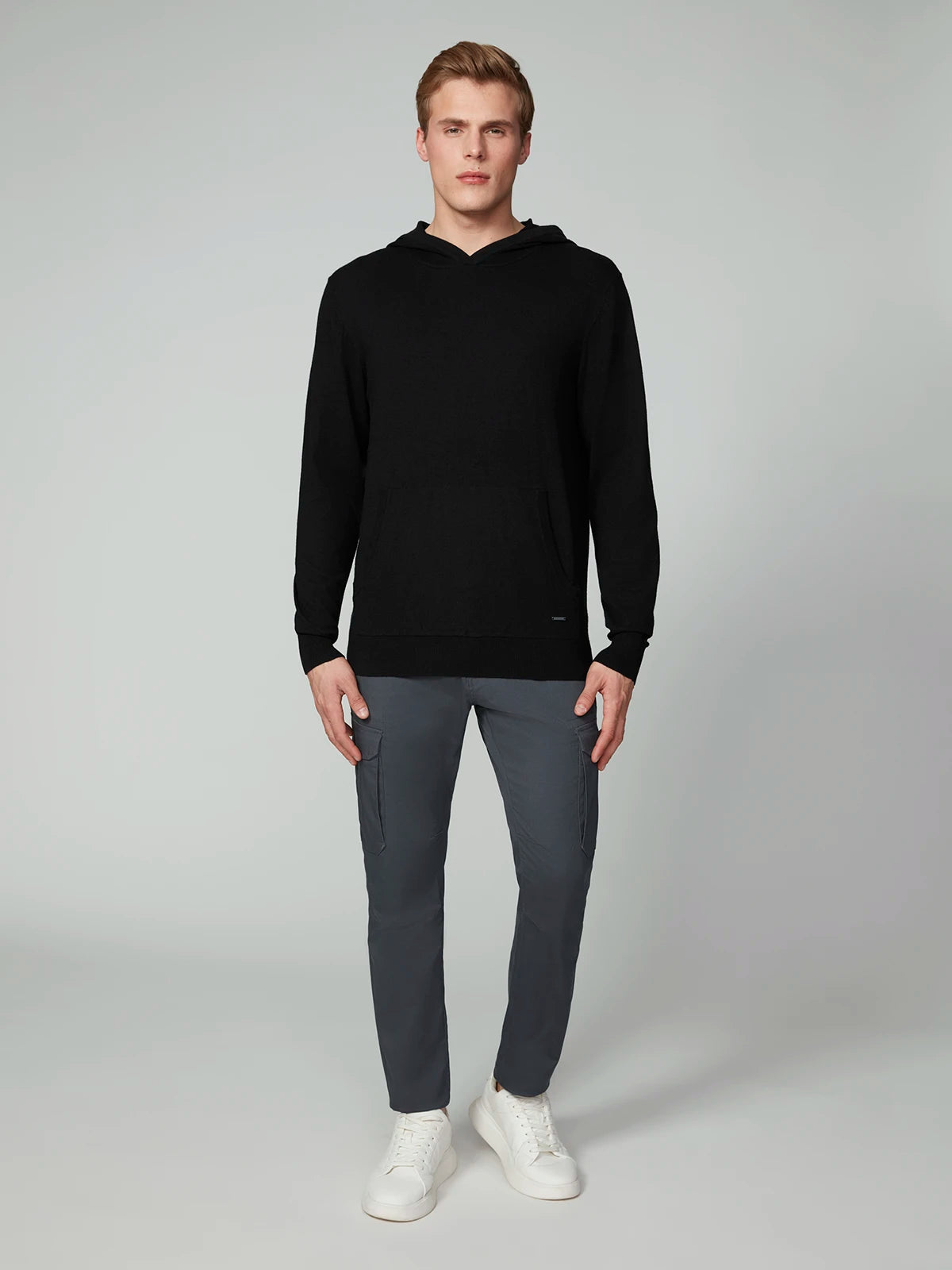 A man stands facing forward in a PROJEK RAW MENS CASHMERE TOUCH HOODIE SWEATER, gray slim-fit pants, and white sneakers against a plain light gray background.