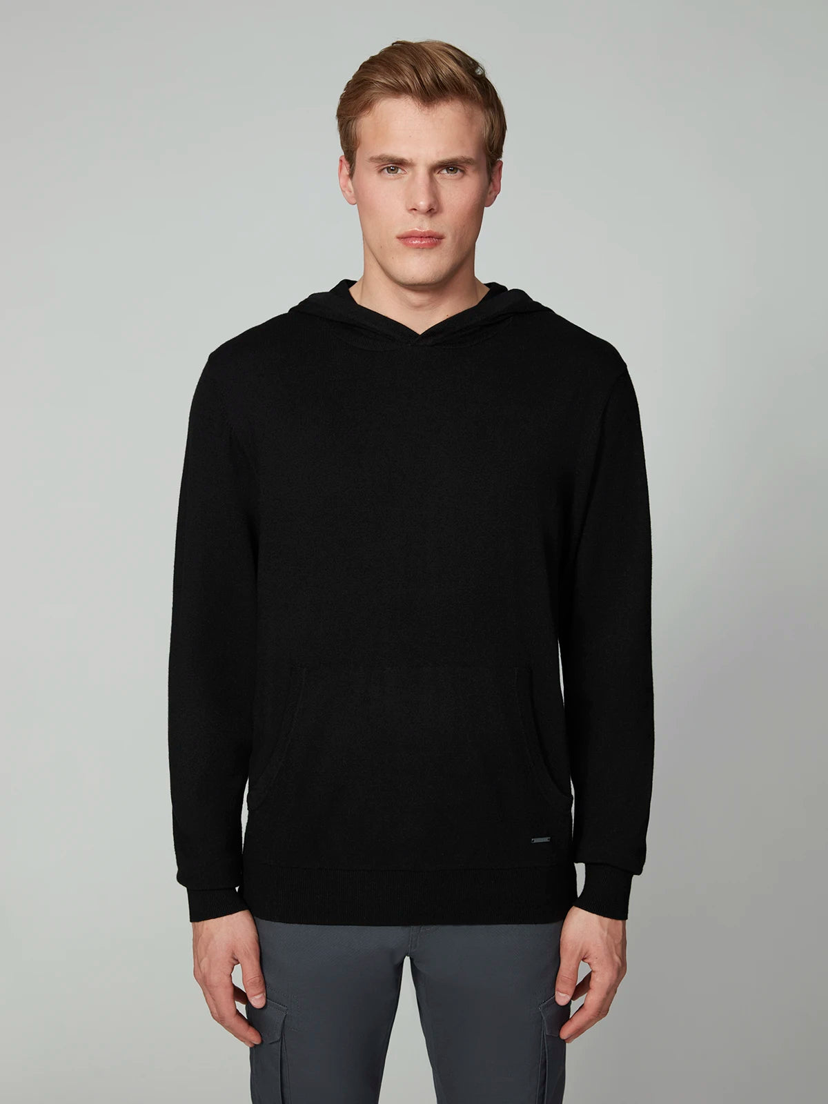 A man with short blond hair wears a black PROJEK RAW MENS CASHMERE TOUCH HOODIE SWEATER and gray pants, standing against a plain light gray background, facing the camera with a neutral expression.