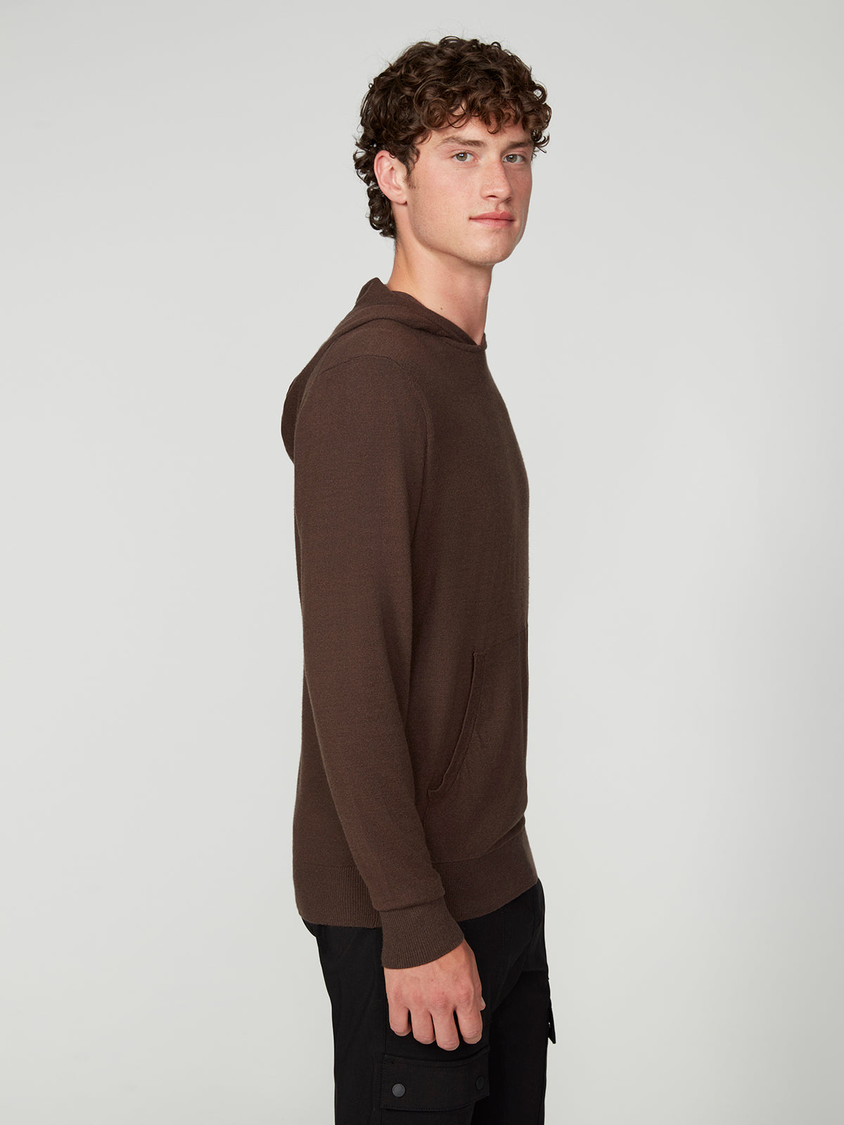 A young man with curly hair stands sideways against a plain light gray background, wearing the PROJEK RAW MENS CASHMERE TOUCH HOODIE SWEATER and black pants.
