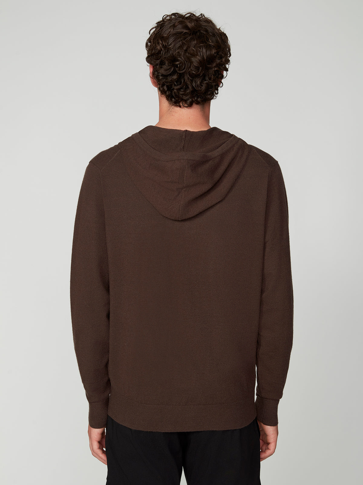 A man with short curly hair is seen from behind, wearing a PROJEK RAW MENS CASHMERE TOUCH HOODIE SWEATER in brown and black pants, standing against a plain light gray background.