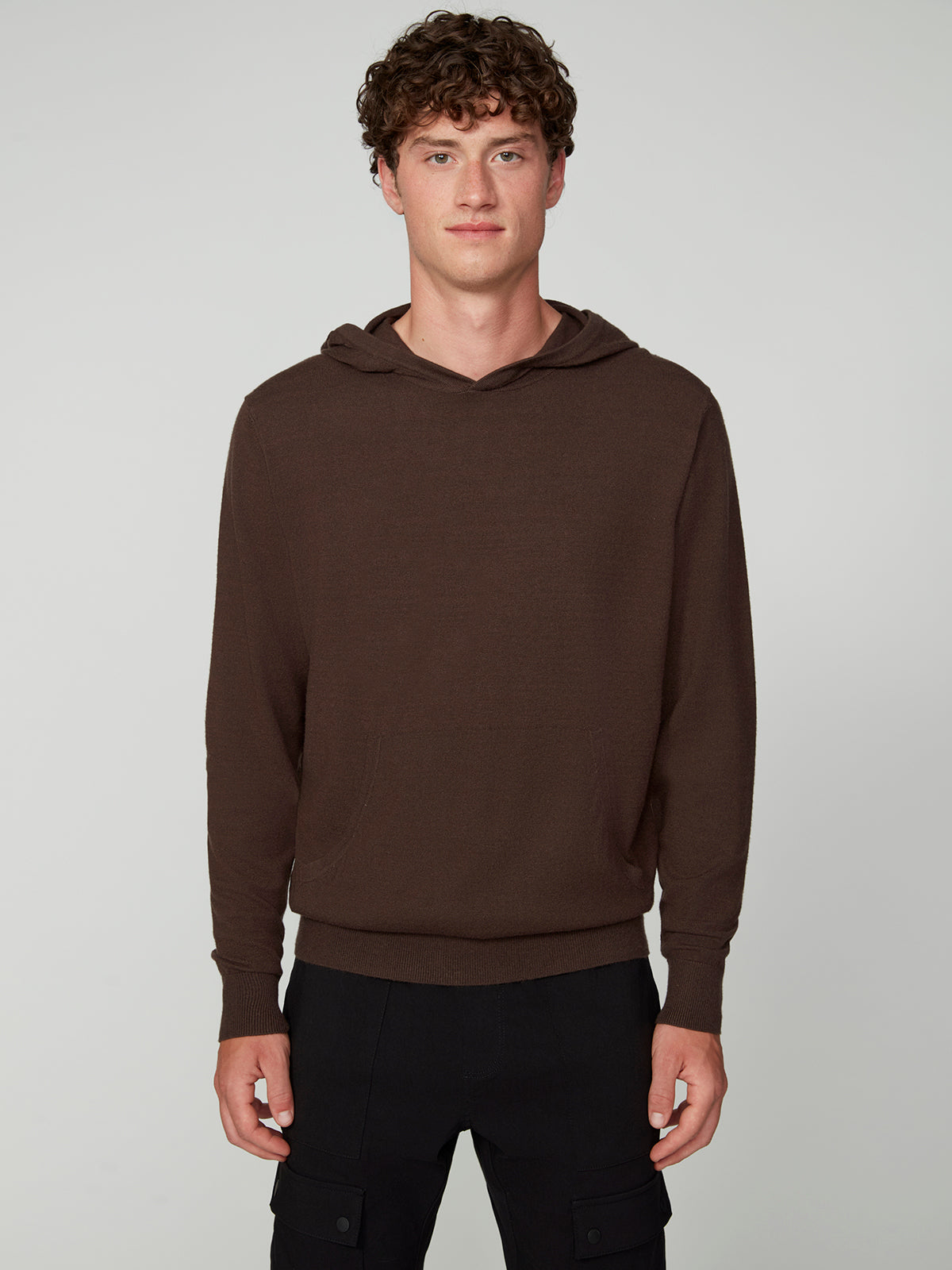 A young man with curly brown hair wears a PROJEK RAW MENS CASHMERE TOUCH HOODIE SWEATER and black pants, standing against a light gray background, facing the camera with a neutral expression.