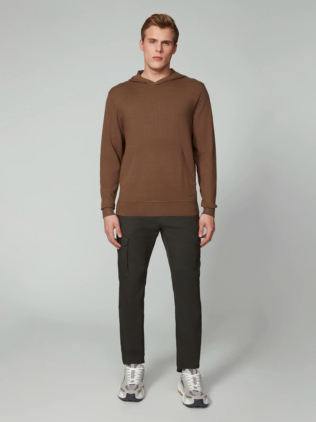 A man faces forward in a PROJEK RAW MENS CASHMERE TOUCH HOODIE SWEATER, paired with dark gray cargo pants and white sneakers against a plain, light backdrop.