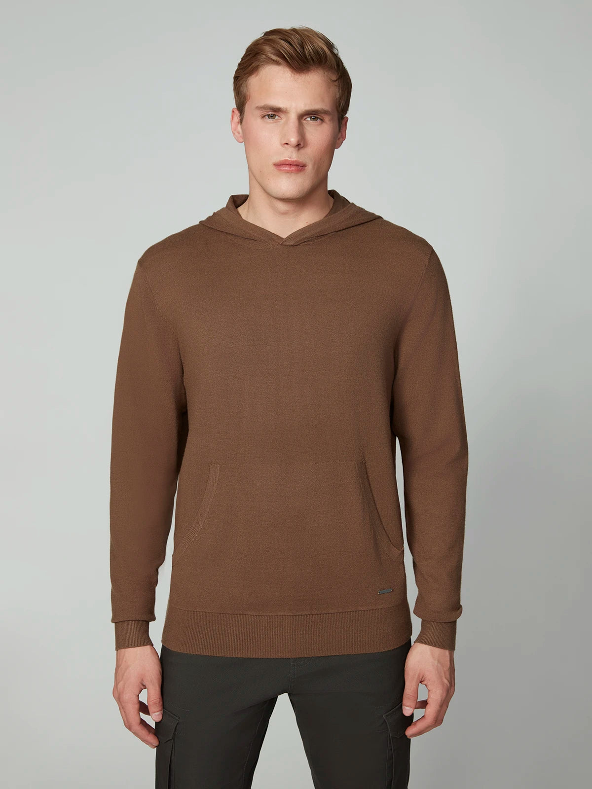 A young man with short light brown hair wears a PROJEK RAW MENS CASHMERE TOUCH HOODIE SWEATER with front pockets and dark pants, standing against a plain light gray background and looking straight at the camera.