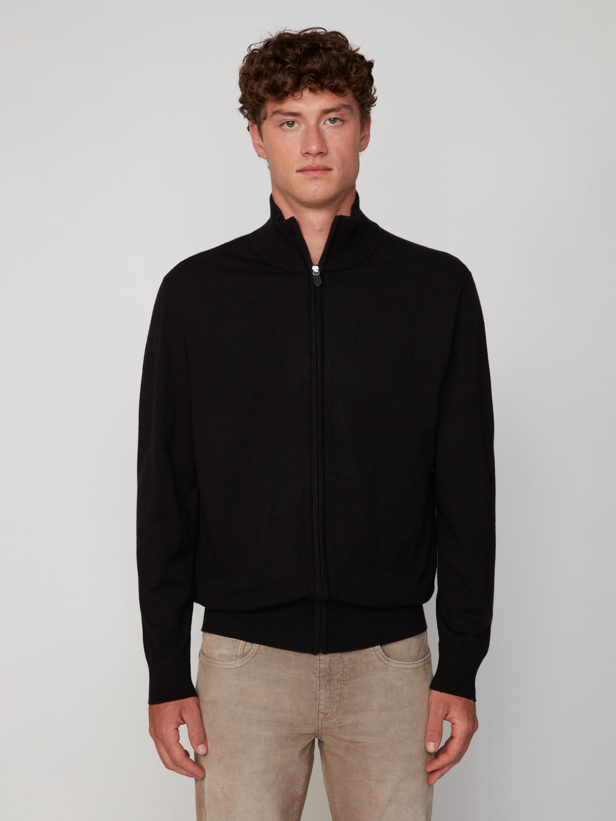A young man with curly brown hair models the PROJEK RAW MENS ZIP-UP CASHMERE TOUCH SWEATER and beige jeans, standing before a plain light gray background and looking straight at the camera with a neutral expression.