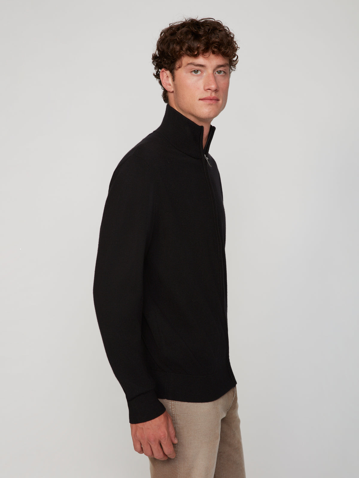 A young man with curly brown hair models the PROJEK RAW MENS ZIP-UP CASHMERE TOUCH SWEATER in black with beige pants, standing sideways and looking at the camera against a plain light background.