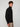A young man with curly brown hair models the PROJEK RAW MENS ZIP-UP CASHMERE TOUCH SWEATER in black with beige pants, standing sideways and looking at the camera against a plain light background.