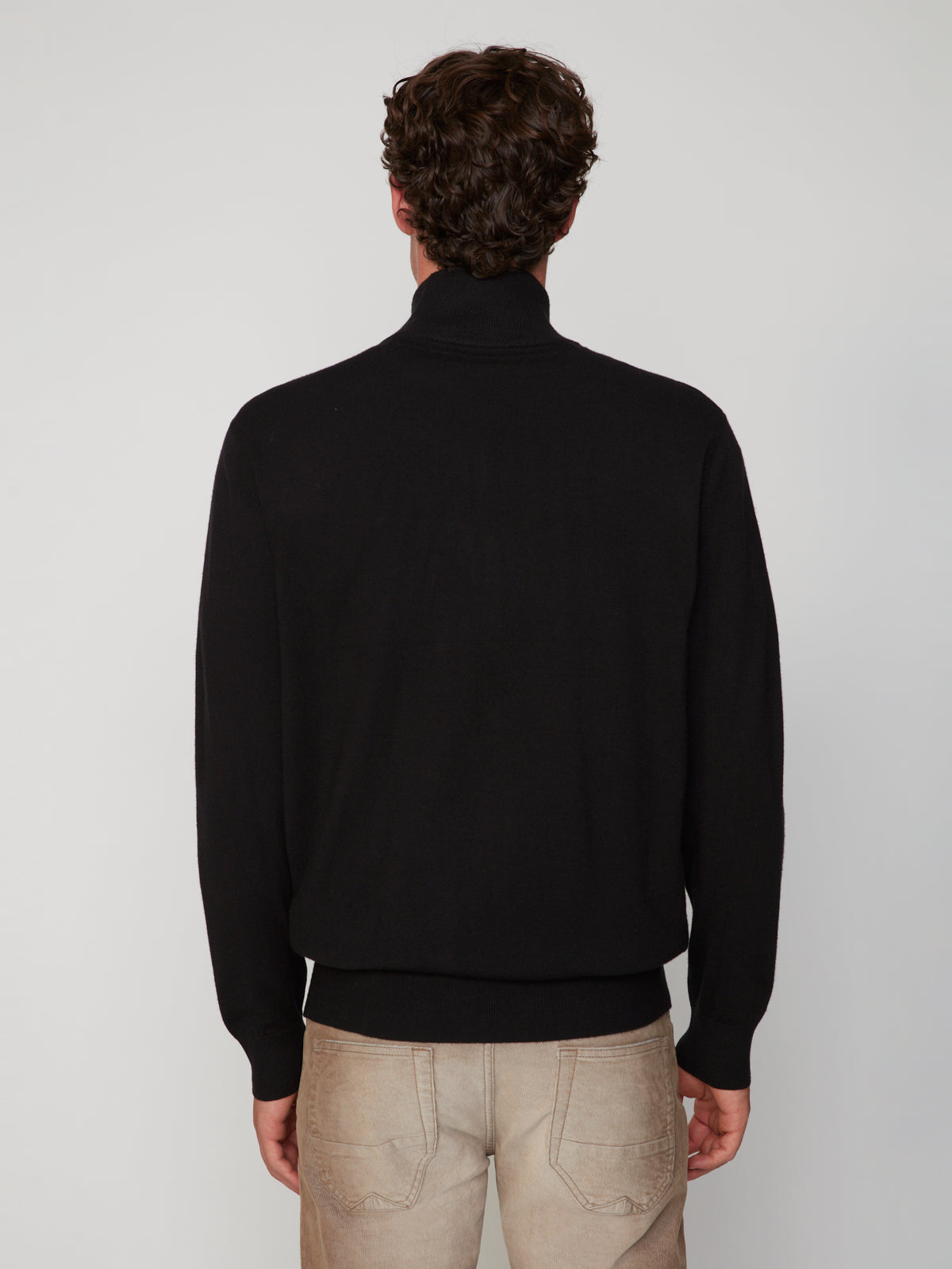 A man with curly brown hair faces away from the camera, wearing a black PROJEK RAW MENS ZIP-UP CASHMERE TOUCH SWEATER and light beige jeans against a plain light background.