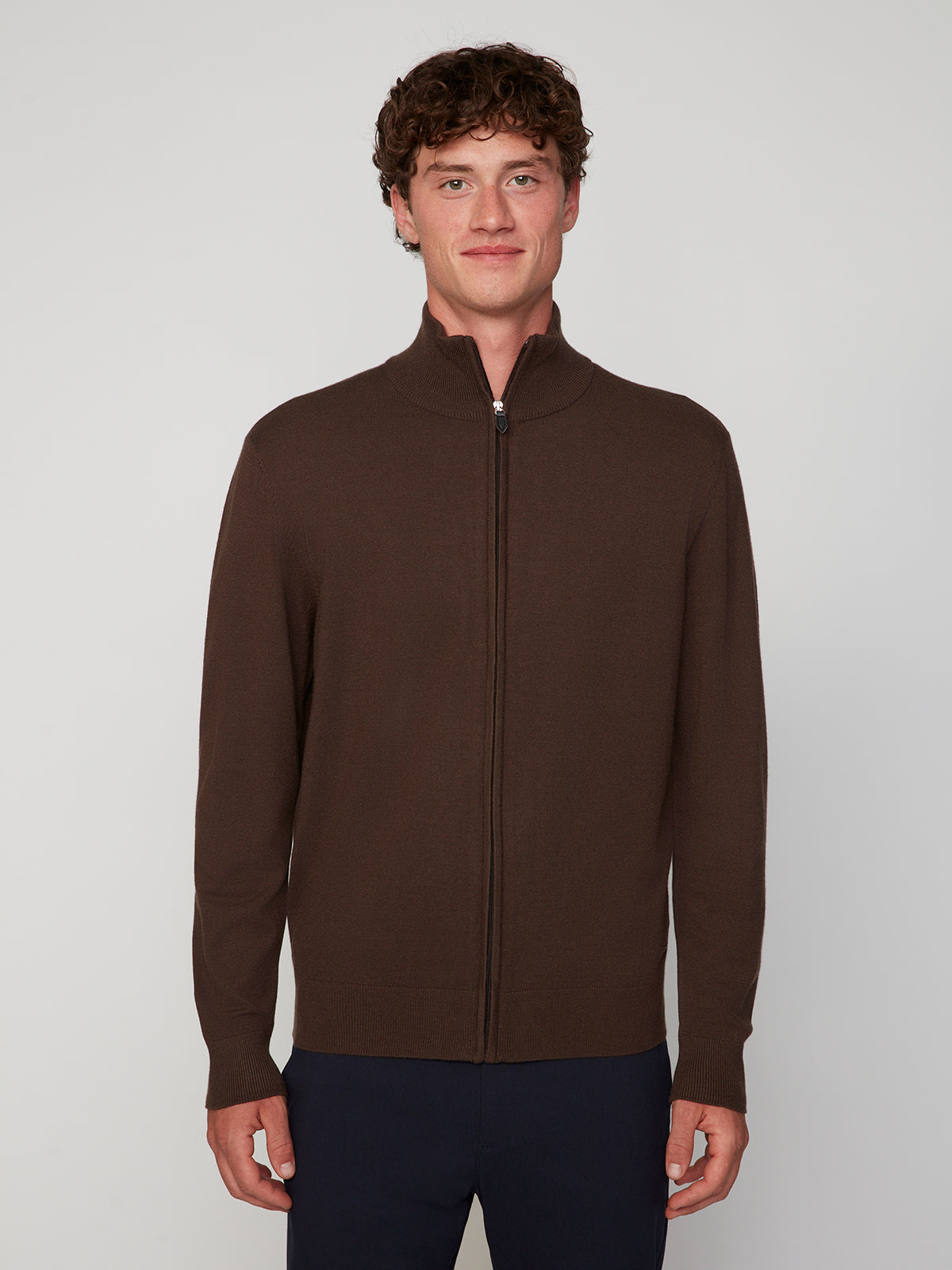 A young man faces the camera wearing a PROJEK RAW MENS ZIP-UP CASHMERE TOUCH SWEATER with a high collar and dark pants, set against a plain light gray background.