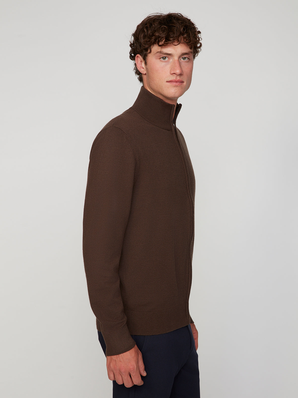 A man with curly brown hair models the PROJEK RAW MENS ZIP-UP CASHMERE TOUCH SWEATER and navy pants, standing sideways against a plain light gray background while looking at the camera.