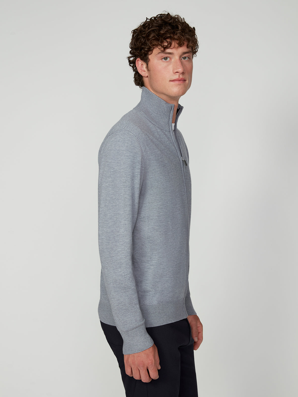 A young man with curly brown hair stands sideways, wearing a light gray PROJEK RAW MENS ZIP-UP CASHMERE TOUCH SWEATER and dark pants, looking at the camera against a plain light background.