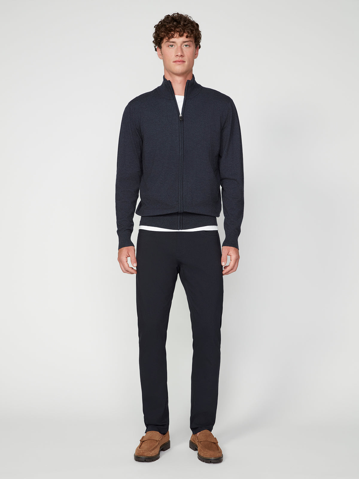 A young man with curly brown hair faces forward, wearing the PROJEK RAW MENS ZIP-UP CASHMERE TOUCH SWEATER over a white shirt, paired with dark pants and brown loafers, standing against a plain light background.