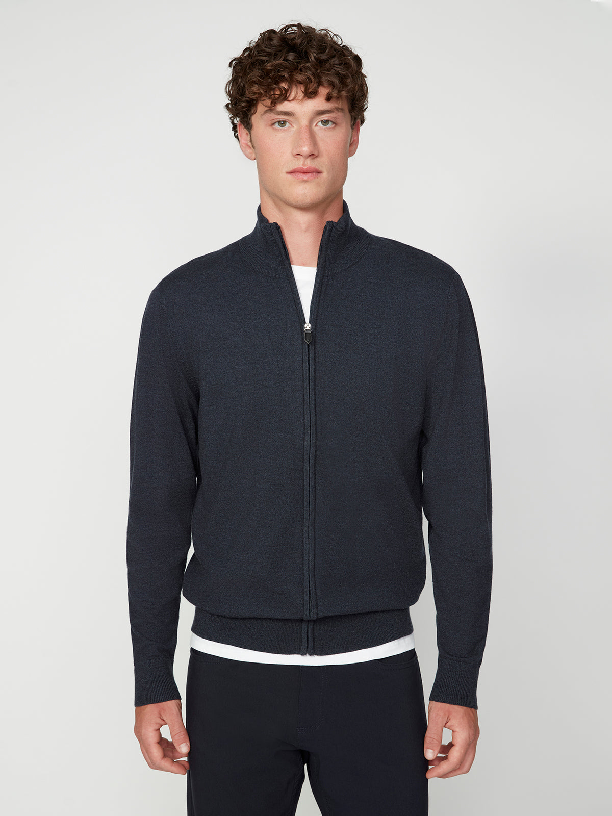 A young man with curly brown hair wears a PROJEK RAW MENS ZIP-UP CASHMERE TOUCH SWEATER over a white t-shirt, standing against a light gray background and looking directly at the camera.
