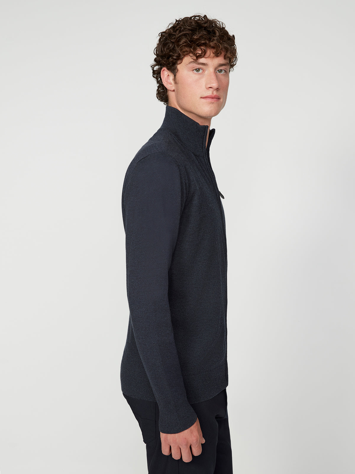 A young man with curly hair stands sideways, facing left, wearing a dark PROJEK RAW MENS ZIP-UP CASHMERE TOUCH SWEATER and dark pants against a plain white background.