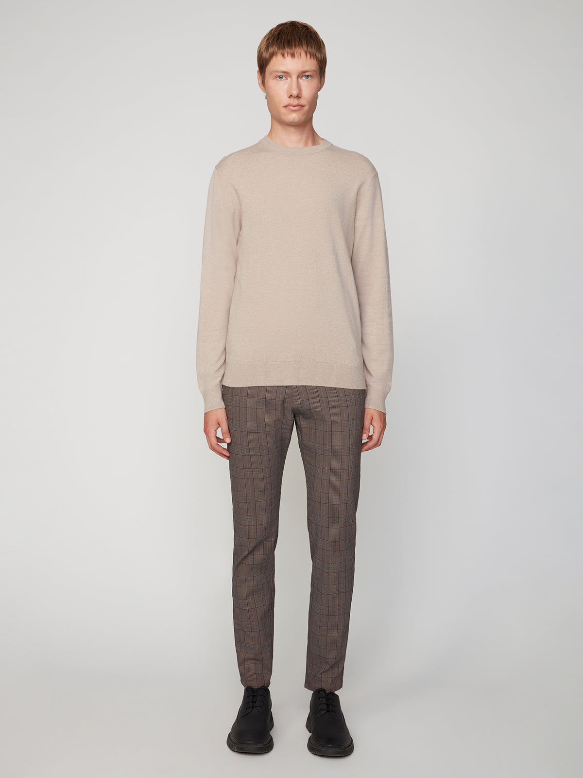 A person stands against a plain light background, wearing a beige PROJEK RAW MENS CREW NECK CASHMERE TOUCH SWEATER, brown plaid trousers, and black shoes.