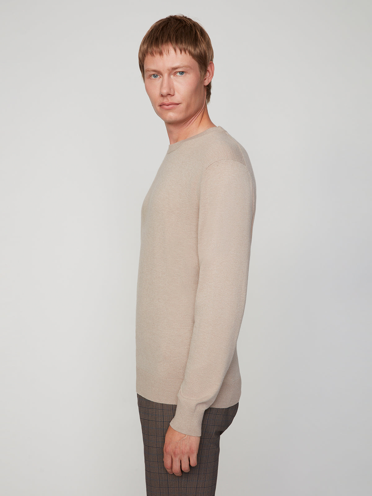 A person with short, light brown hair wears the PROJEK RAW MENS CREW NECK CASHMERE TOUCH SWEATER in beige and plaid pants, posing sideways and looking at the camera against a plain light background.
