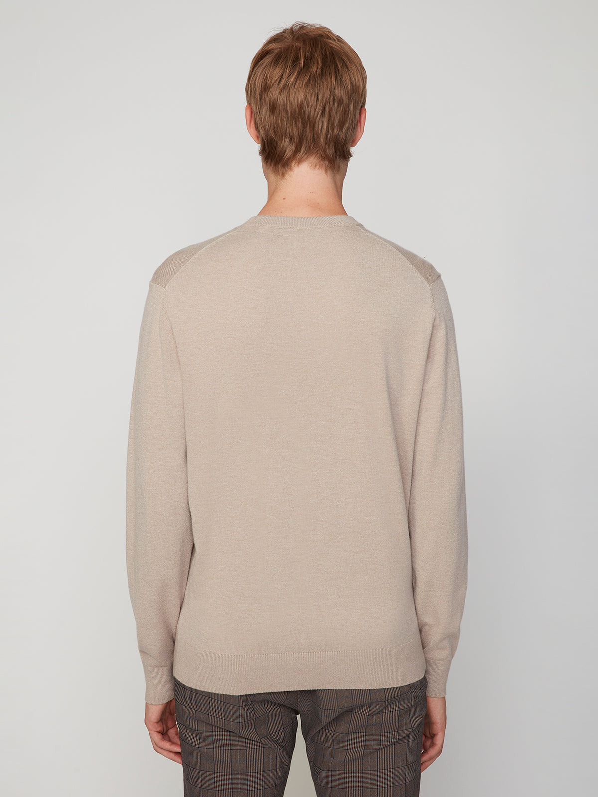 A person with short light brown hair is seen from behind wearing a beige PROJEK RAW MENS CREW NECK CASHMERE TOUCH SWEATER and brown plaid pants, standing against a plain light gray background.