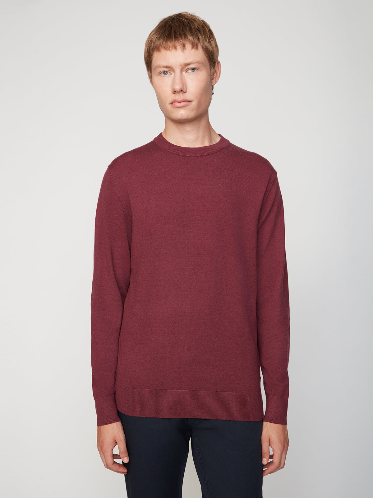A man with short light brown hair wears a PROJEK RAW MENS CREW NECK CASHMERE TOUCH SWEATER in maroon and dark pants, standing against a plain light gray background, looking directly at the camera with a neutral expression.