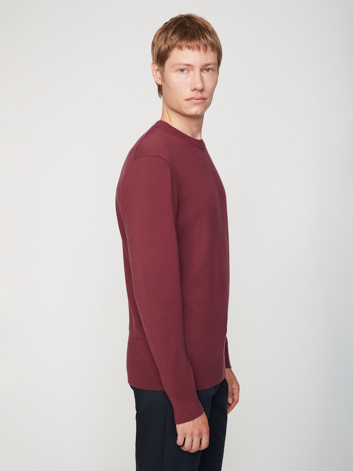 A man with short light brown hair models the PROJEK RAW MENS CREW NECK CASHMERE TOUCH SWEATER in maroon with dark pants, standing sideways and looking at the camera against a plain light background.