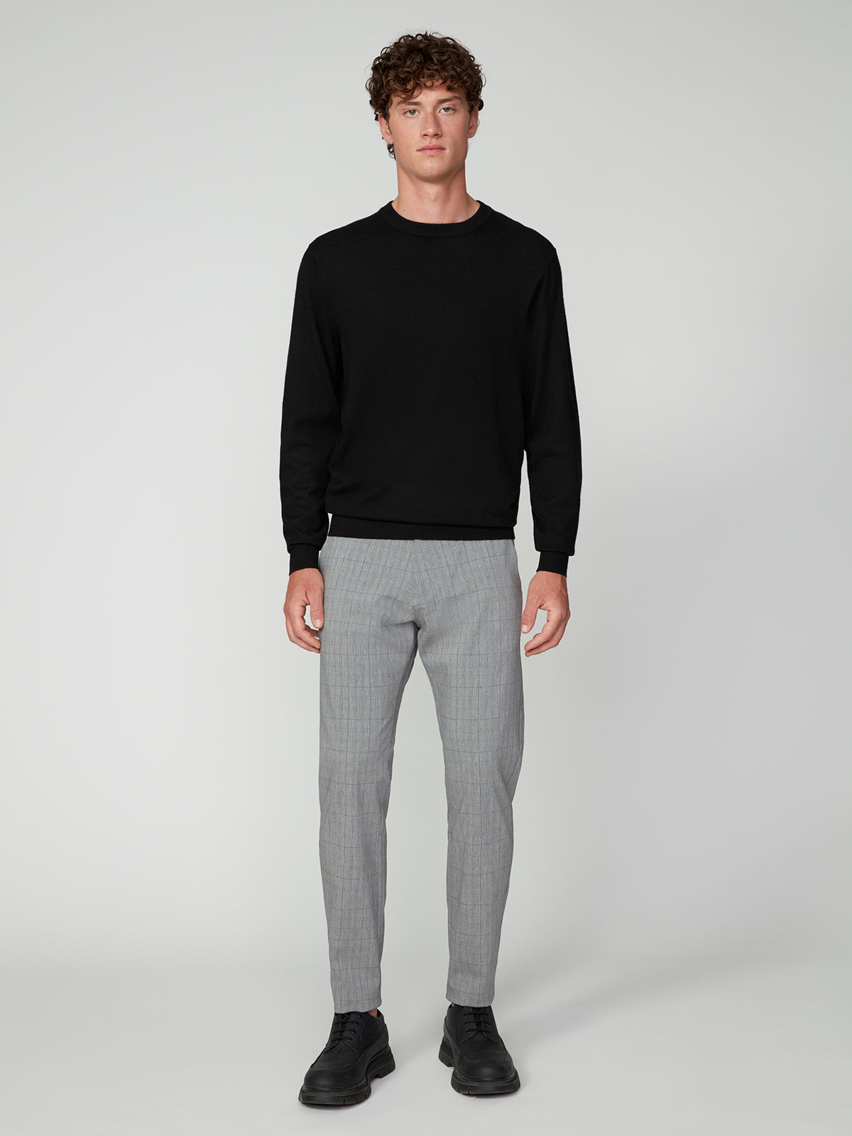 A young man with curly hair faces forward, wearing a PROJEK RAW MENS CREW NECK CASHMERE TOUCH SWEATER in black, paired with light gray plaid pants and black shoes against a plain, light-colored background.