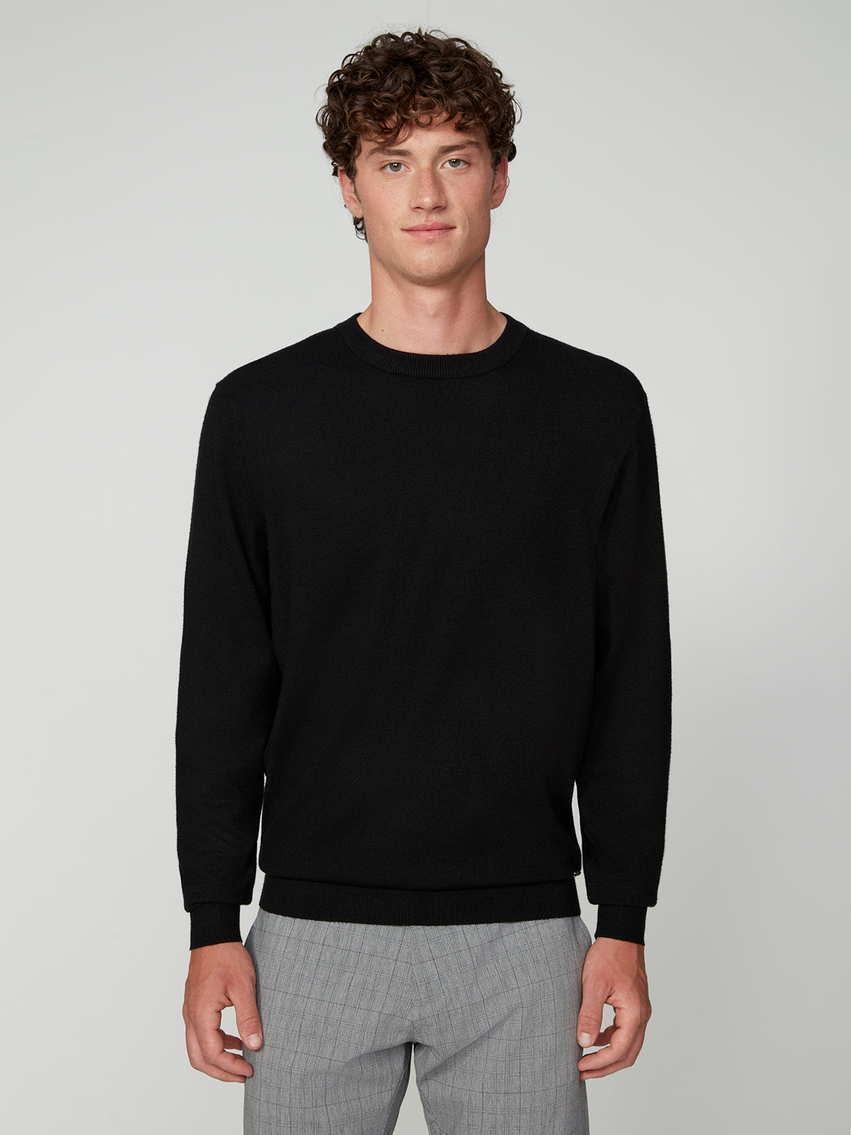 A young man with curly brown hair wears a PROJEK RAW MENS CREW NECK CASHMERE TOUCH SWEATER and light gray plaid pants, standing against a plain light gray background, looking at the camera with a neutral expression.